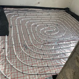 Hydro-Plumbing Ltd - Białe rury ogrzewania podłogowego ułożone spiralnie na folii z czerwoną kratką, przymocowane czarnymi uchwytami, widok z góry, przygotowane do zalania wylewką.