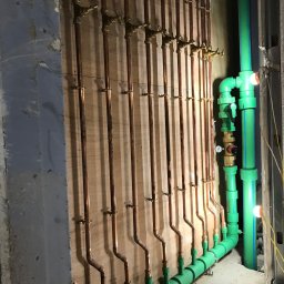 Hydro-Plumbing Ltd - Miedziane i zielone rury instalacji wodnej zamontowane na ścianie z drewnianą płytą. Widoczne liczne zawory i złączki. Część instalacji ukryta w ścianie.