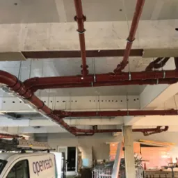 Czerwone rury instalacji podwieszone pod stropem budynku w trakcie budowy, widoczne mocowania i elementy złączne. Częściowo widoczny samoch&oacute;d firmy Openreach.