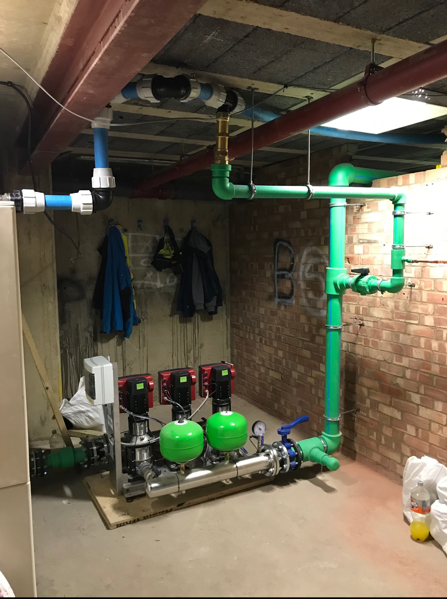 Kompleksowa instalacja hydrauliczna z zielonymi rurami i czerwonymi pompami w kotłowni, widoczne elementy przyłączeniowe i osprzęt. Ściana z cegły z graffiti.