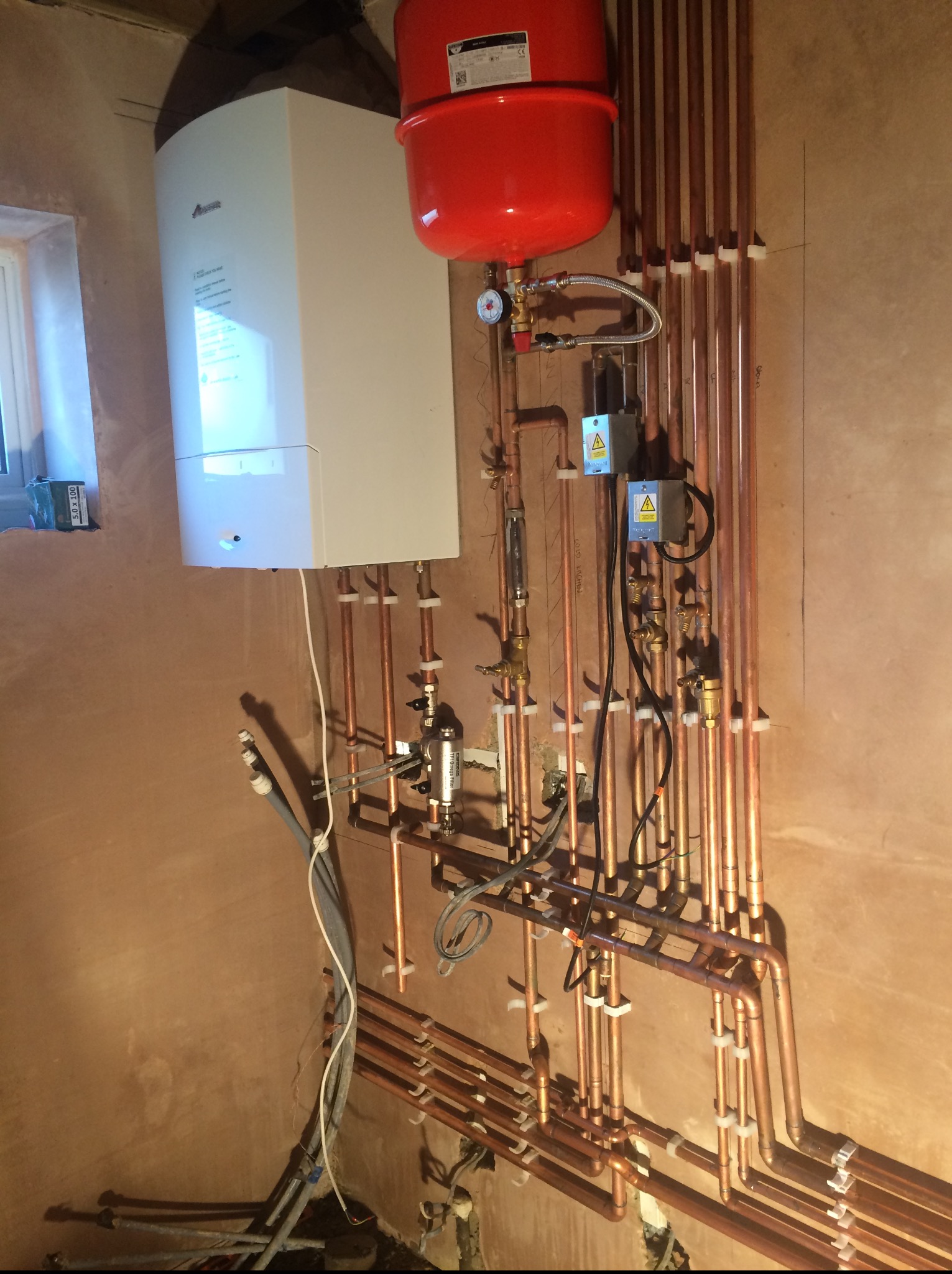 Nowoczesny piec gazowy z czerwonym zbiornikiem wyrównawczym i systemem rur miedzianych, zamontowany na ścianie w trakcie instalacji. Widoczne zawory i elementy sterujące.