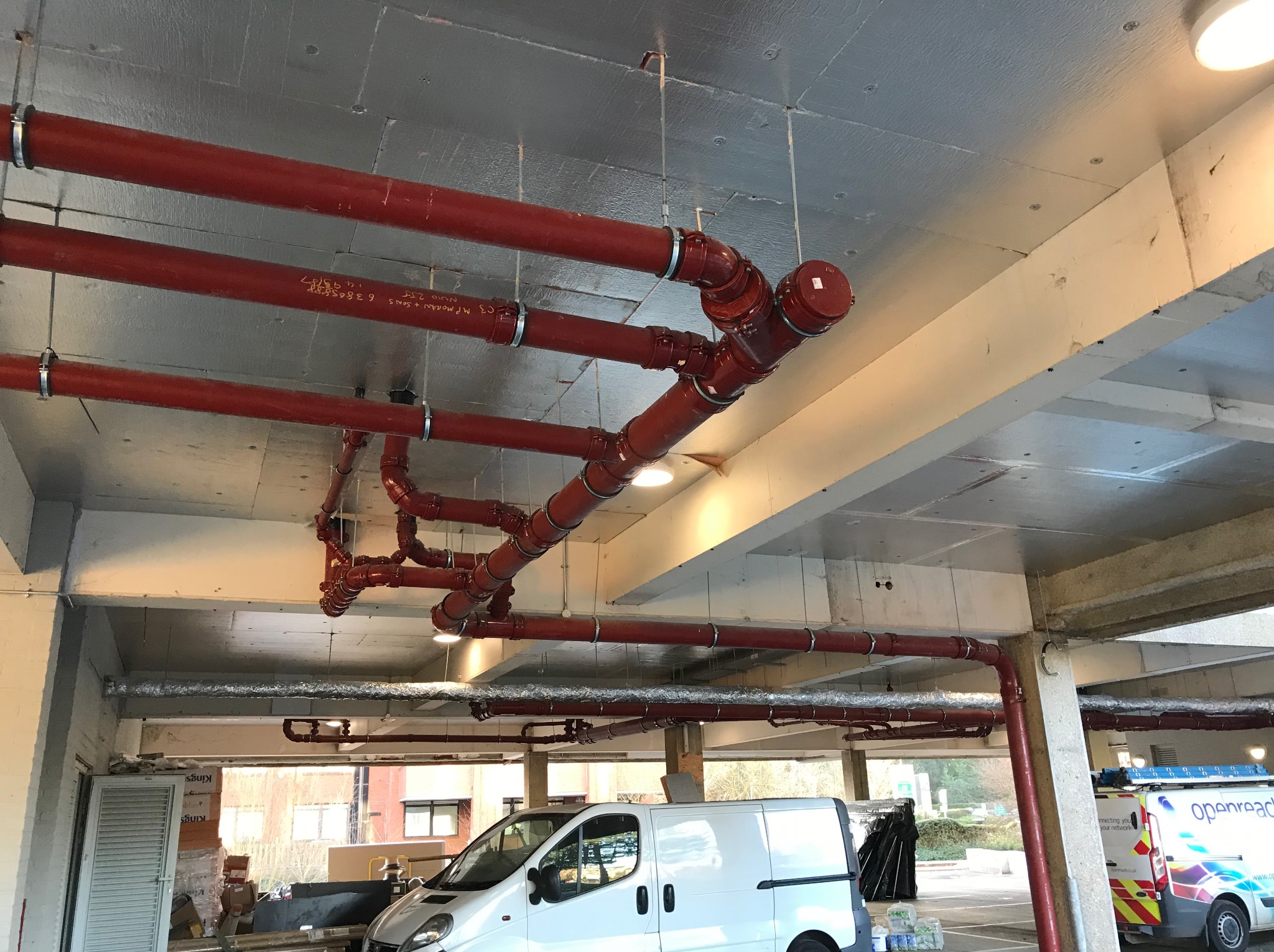 Czerwone rury instalacji przeciwpożarowej, zamontowane pod sufitem parkingu, z widocznymi złączkami i uchwytami. W tle zaparkowane busy, w tym jeden z logo firmy.