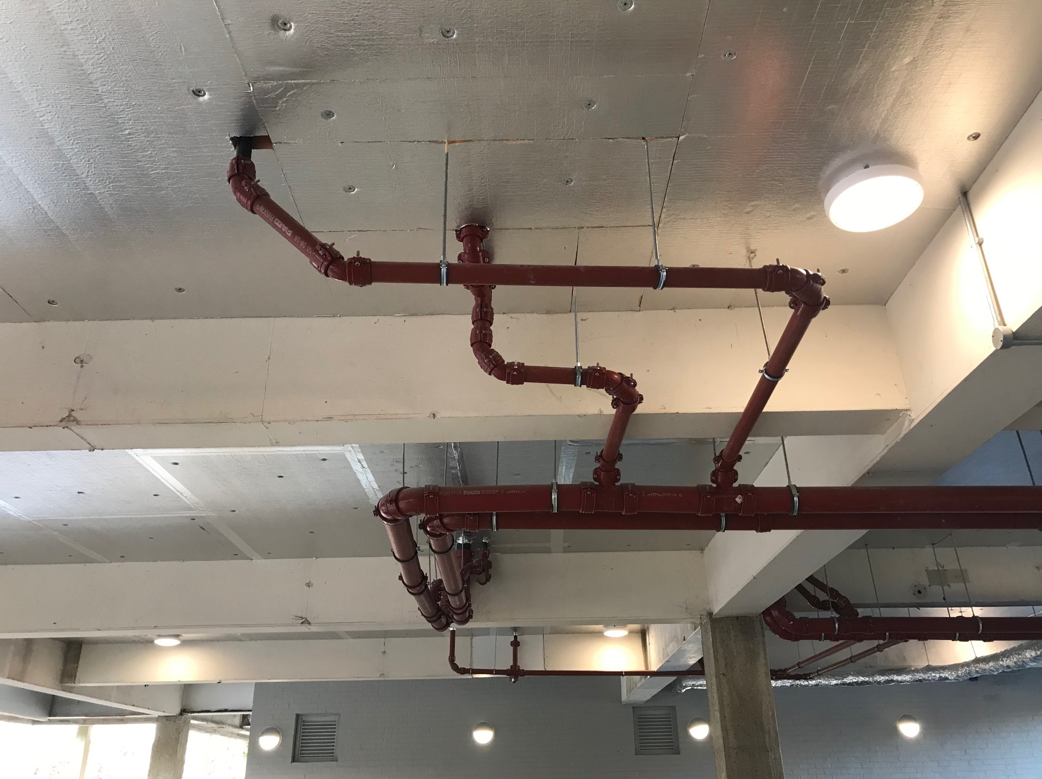 Czerwone rury instalacji hydraulicznej pod sufitem, widoczne mocowania i łączenia, metaliczne wykończenie sufitu, oświetlenie sufitowe.