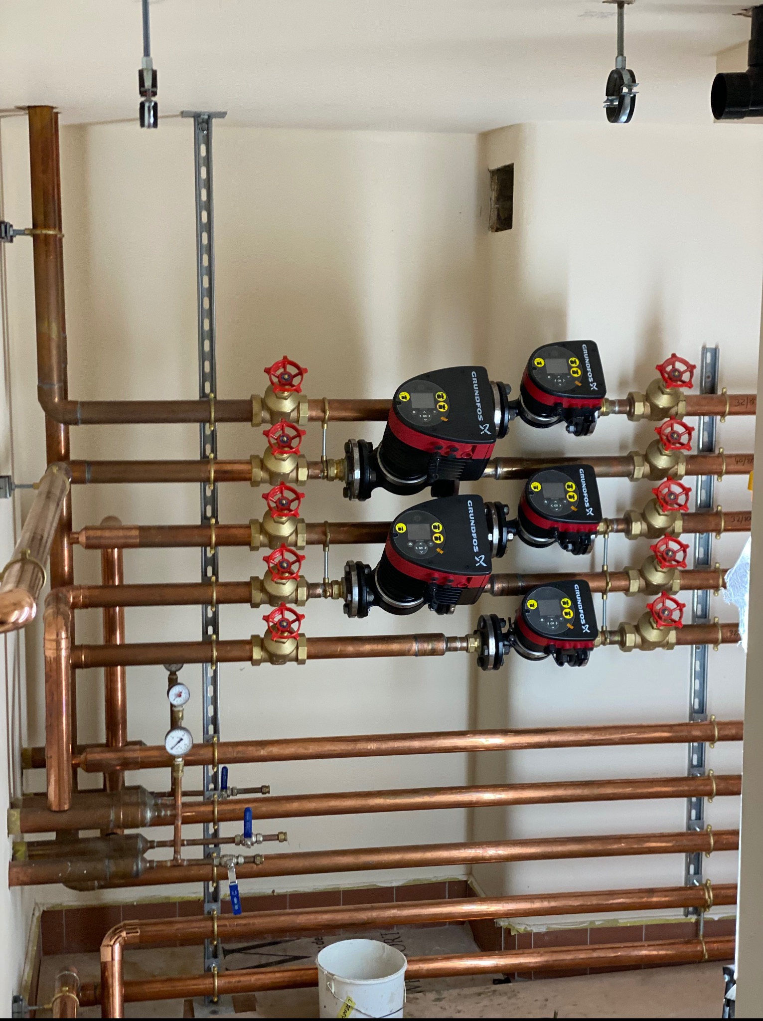 Miedziana instalacja grzewcza z zaworami i pompami obiegowymi Grundfos, zamontowana na ścianie. Widoczne elementy mocujące i manometry.