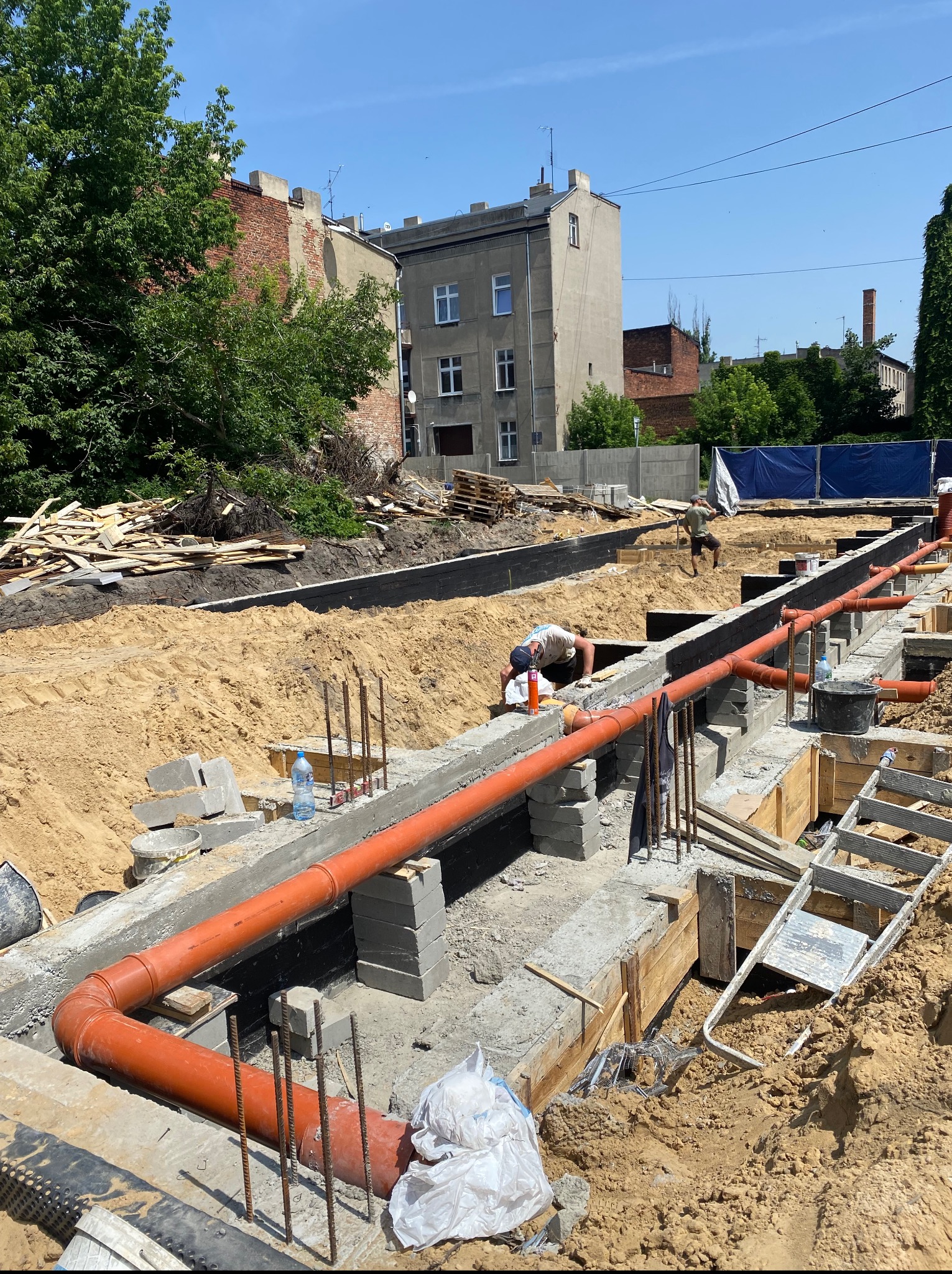 Montaż pomarańczowych rur kanalizacyjnych w wykopie fundamentowym, widoczny pracownik przy łączeniu, częściowo zalane fundamenty, teren budowy z drewnem i piaskiem.