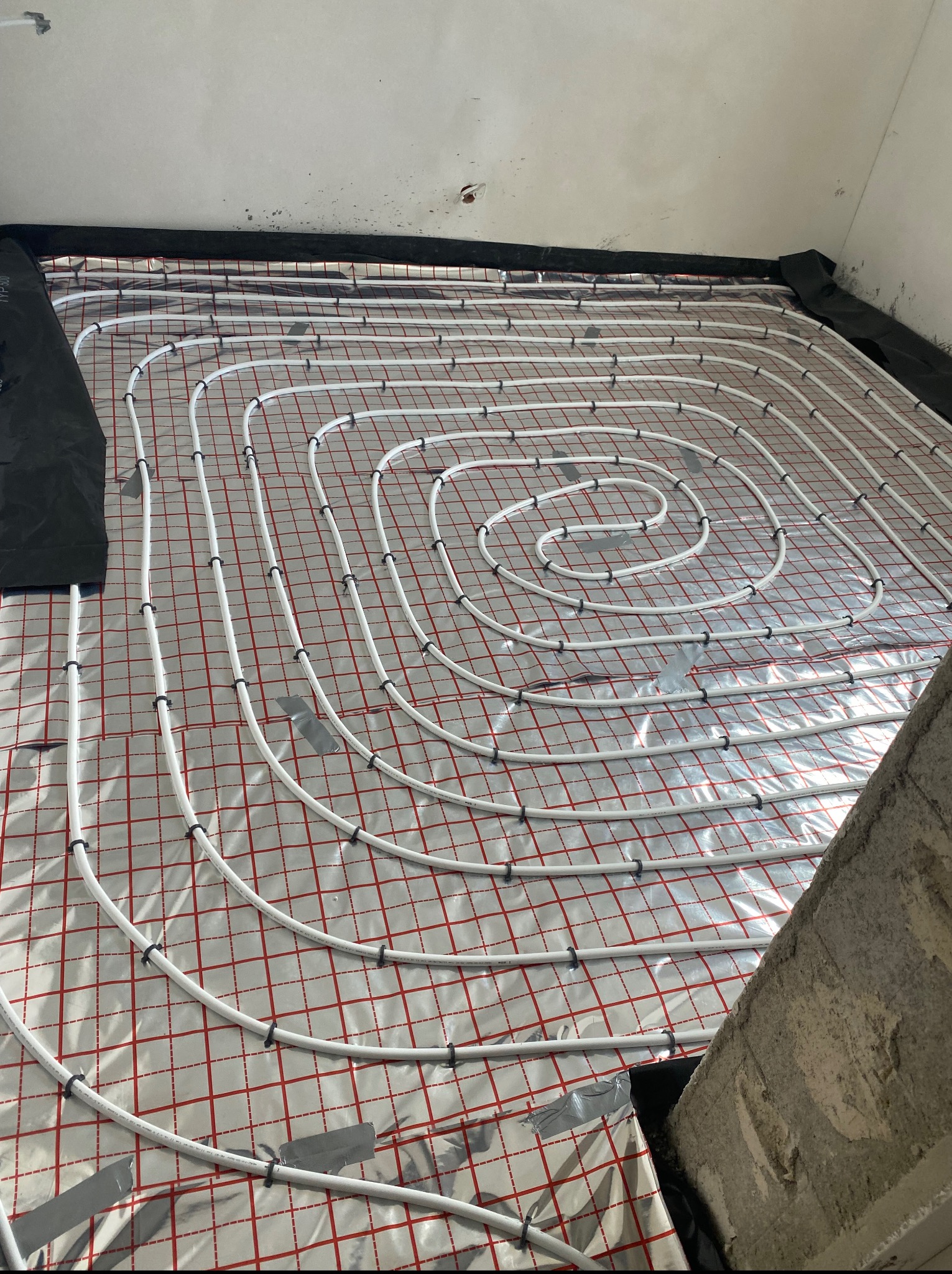 Białe rury ogrzewania podłogowego ułożone spiralnie na folii z czerwoną kratką, przymocowane czarnymi uchwytami, widok z góry, przygotowane do zalania wylewką.