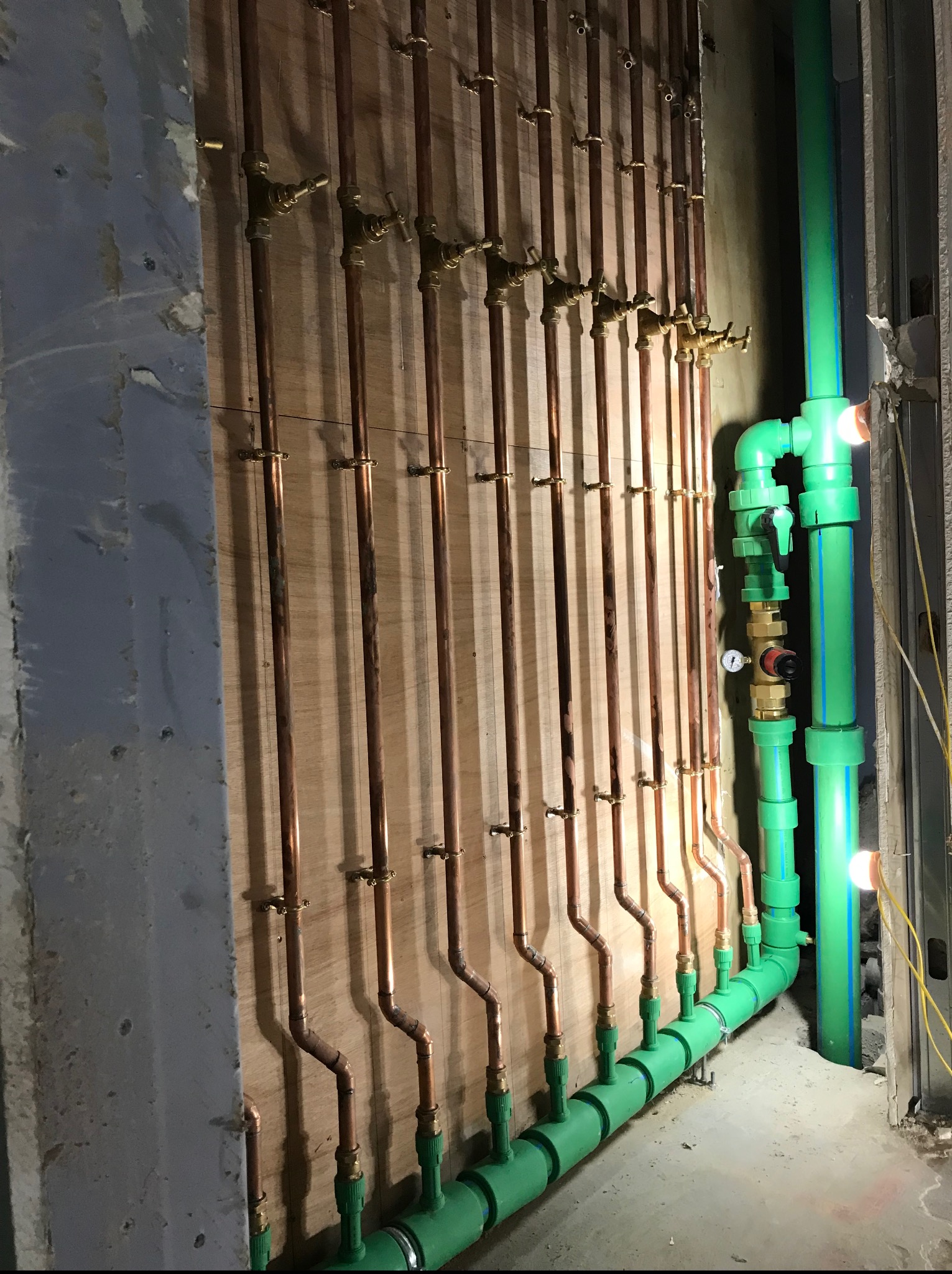 Miedziane i zielone rury instalacji wodnej zamontowane na ścianie z drewnianą płytą. Widoczne liczne zawory i złączki. Część instalacji ukryta w ścianie.