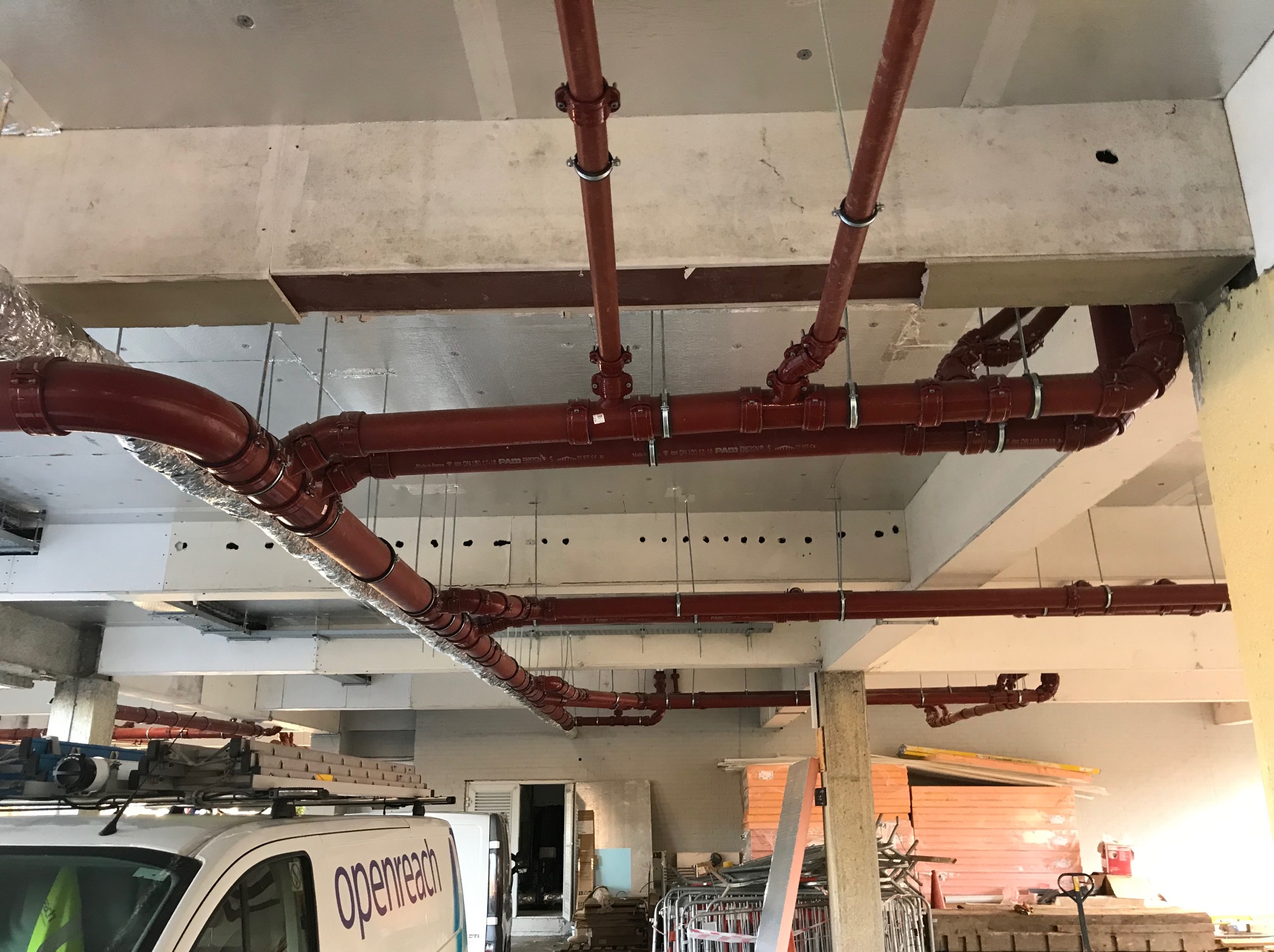 Czerwone rury instalacji podwieszone pod stropem budynku w trakcie budowy, widoczne mocowania i elementy złączne. Częściowo widoczny samochód firmy Openreach.