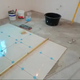 Układanie jasnych płytek podłogowych z użyciem systemu poziomowania; widoczna poziomica, wiadro, puszki z klejem, szpachelka i listwa wykończeniowa.