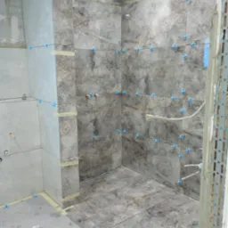 Łazienka w trakcie wykańczania: szare płytki imitujące marmur na ścianach i podłodze, niebieskie krzyżyki dystansowe, widoczne instalacje. Prace glazurnicze w toku.
