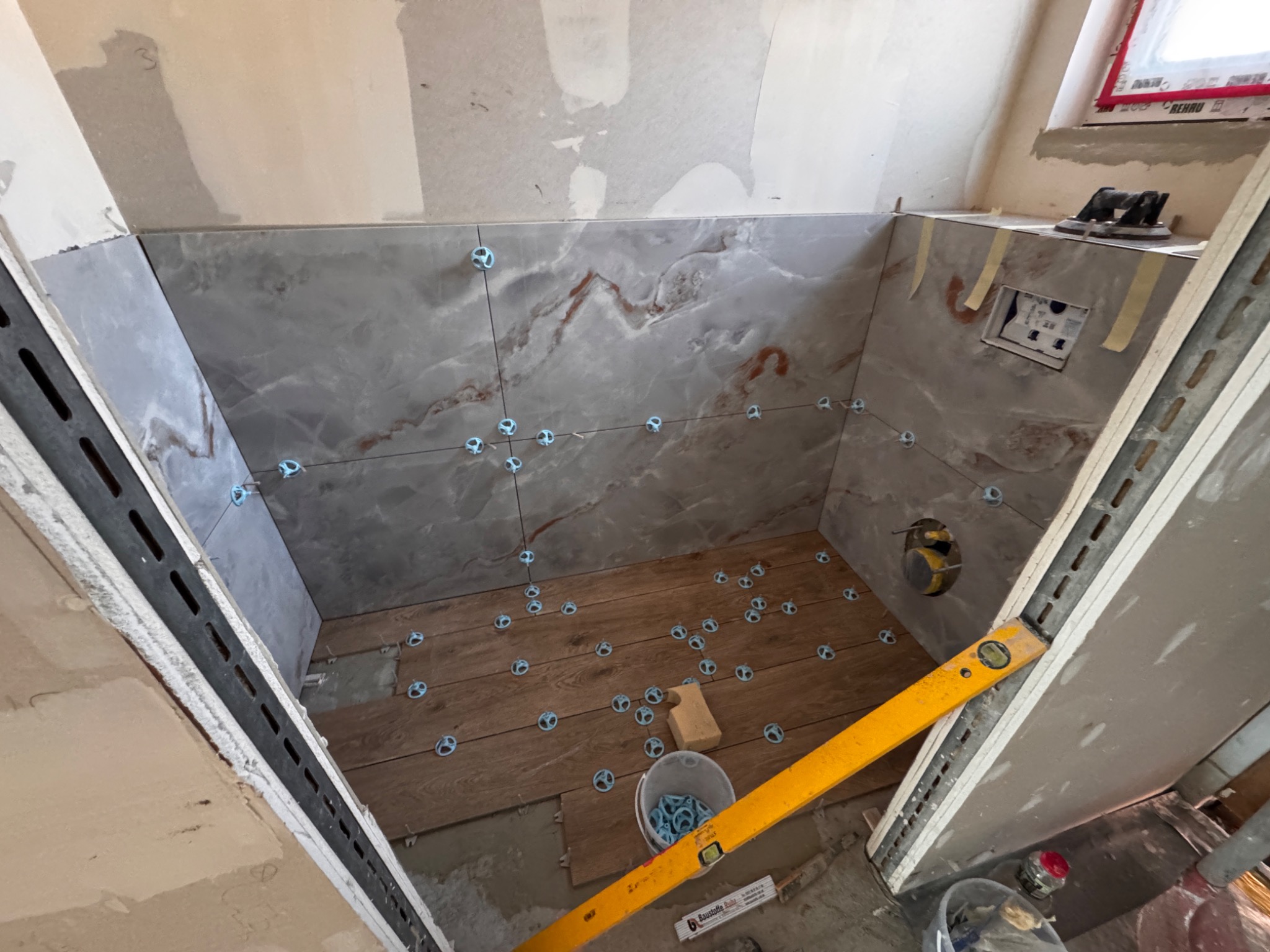 Wnętrze łazienki w trakcie wykańczania: szare, marmurkowe płytki na ścianach z niebieskimi krzyżykami dystansowymi, drewnopodobne płytki na podłodze, poziomica.