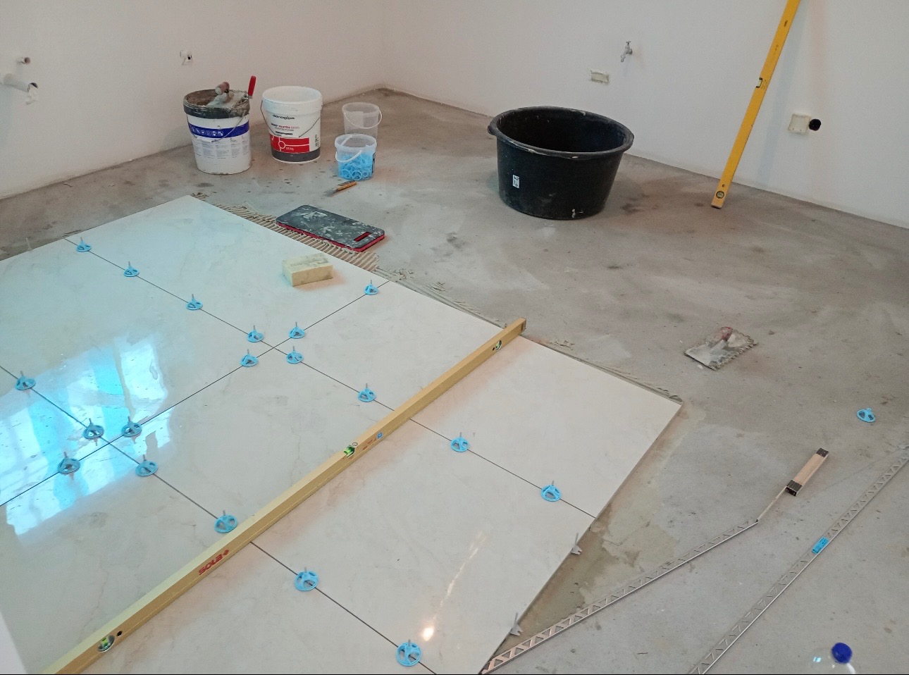 Układanie jasnych płytek podłogowych z użyciem systemu poziomowania; widoczna poziomica, wiadro, puszki z klejem, szpachelka i listwa wykończeniowa.