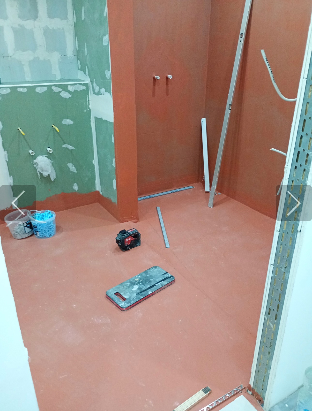 Łazienka w trakcie hydroizolacji: czerwona powłoka na ścianach i podłodze, poziomica, laser krzyżowy i akcesoria glazurnicze. Ściany częściowo pokryte płytami gipsowo-kartonowymi.
