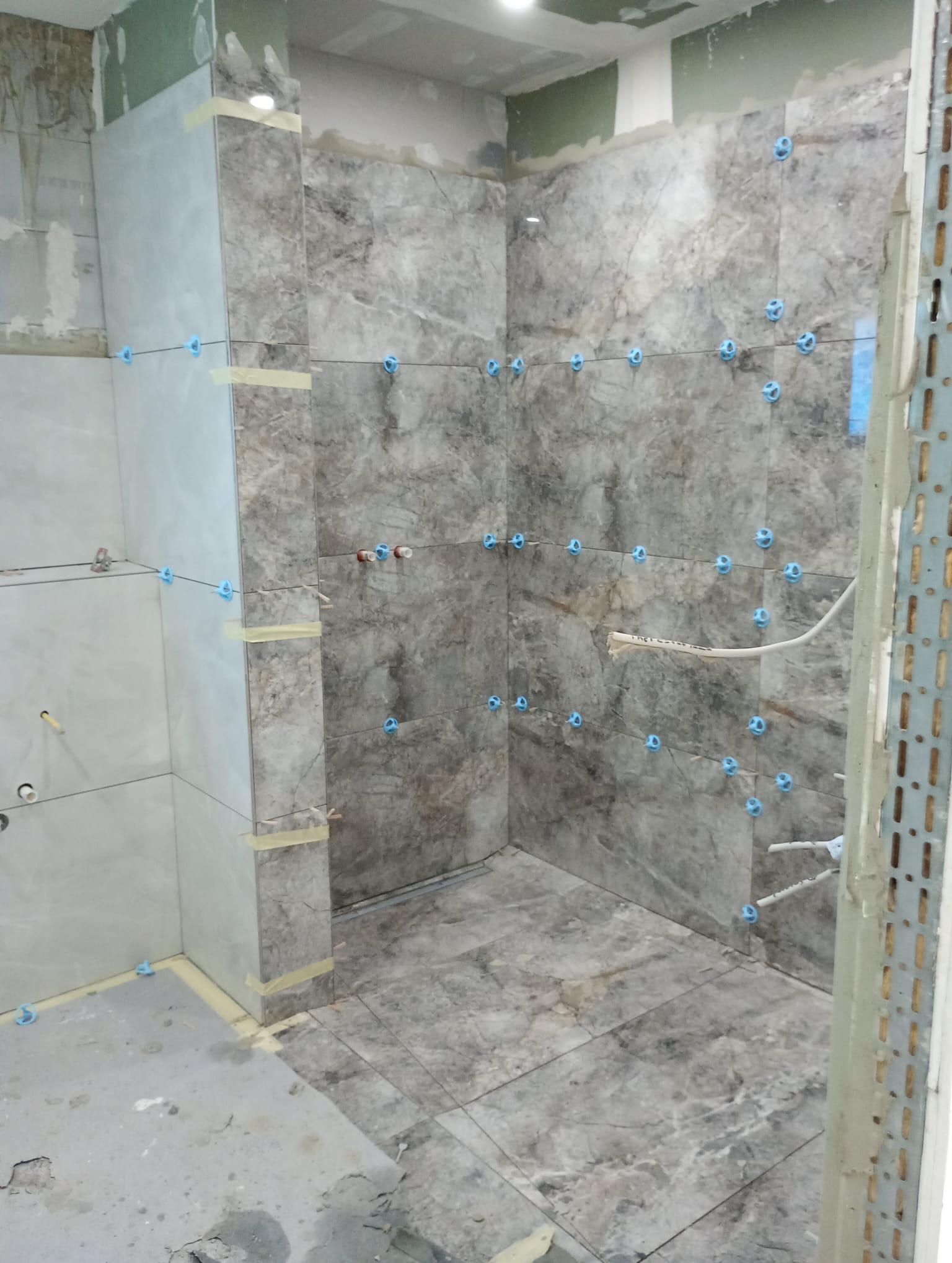 Łazienka w trakcie wykańczania: szare płytki imitujące marmur na ścianach i podłodze, niebieskie krzyżyki dystansowe, widoczne instalacje. Prace glazurnicze w toku.