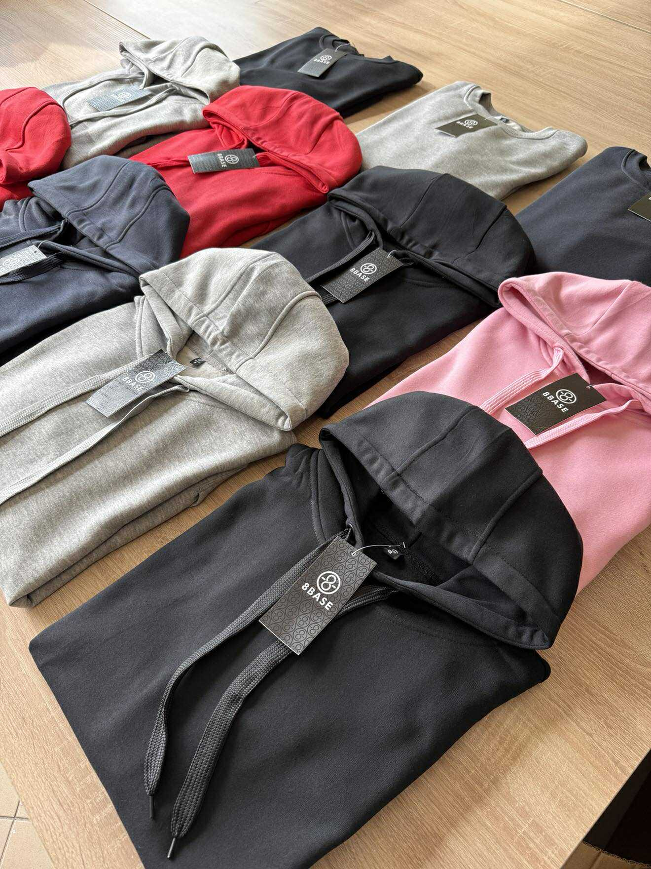 Różnokolorowe bluzy z kapturem marki 8Base ułożone na drewnianym stole. Widoczne metki. Czarny, szary, czerwony, granatowy i różowy. Ujęcie z góry, ostre światło.