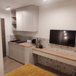 MICHAŁ ZDERSKI EMZZE - Funkcjonalna kuchnia na wymiar w małym mieszkaniu: jasne szafki, blat z drewna, złota armatura, mozaika na ścianie i telewizor. Kompaktowe rozwiązanie.