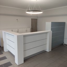 MICHAŁ ZDERSKI EMZZE - Biała lada recepcyjna z poziomymi panelami, oświetlona nowoczesną lampą. W tle szafki zamykane na klucz i szara szafa. Całość w minimalistycznym, nowoczesnym stylu.