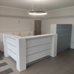 Biała lada recepcyjna z poziomymi panelami, oświetlona nowoczesną lampą. W tle szafki zamykane na klucz i szara szafa. Całość w minimalistycznym, nowoczesnym stylu.