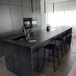 MICHAŁ ZDERSKI EMZZE - Nowoczesna kuchnia z wyspą i hokerami. Ciemnoszara zabudowa meblowa, minimalistyczne lampy wiszące. Na blacie czajnik i akcesoria kuchenne. Prosty, elegancki design.