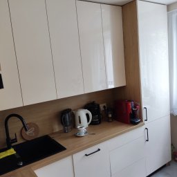 MICHAŁ ZDERSKI EMZZE - Biało-kremowa kuchnia z połyskiem i drewnianym blatem. Widoczny czarny zlew, ekspresy do kawy i czarne, podłużne uchwyty. Górne szafki sięgają sufitu.