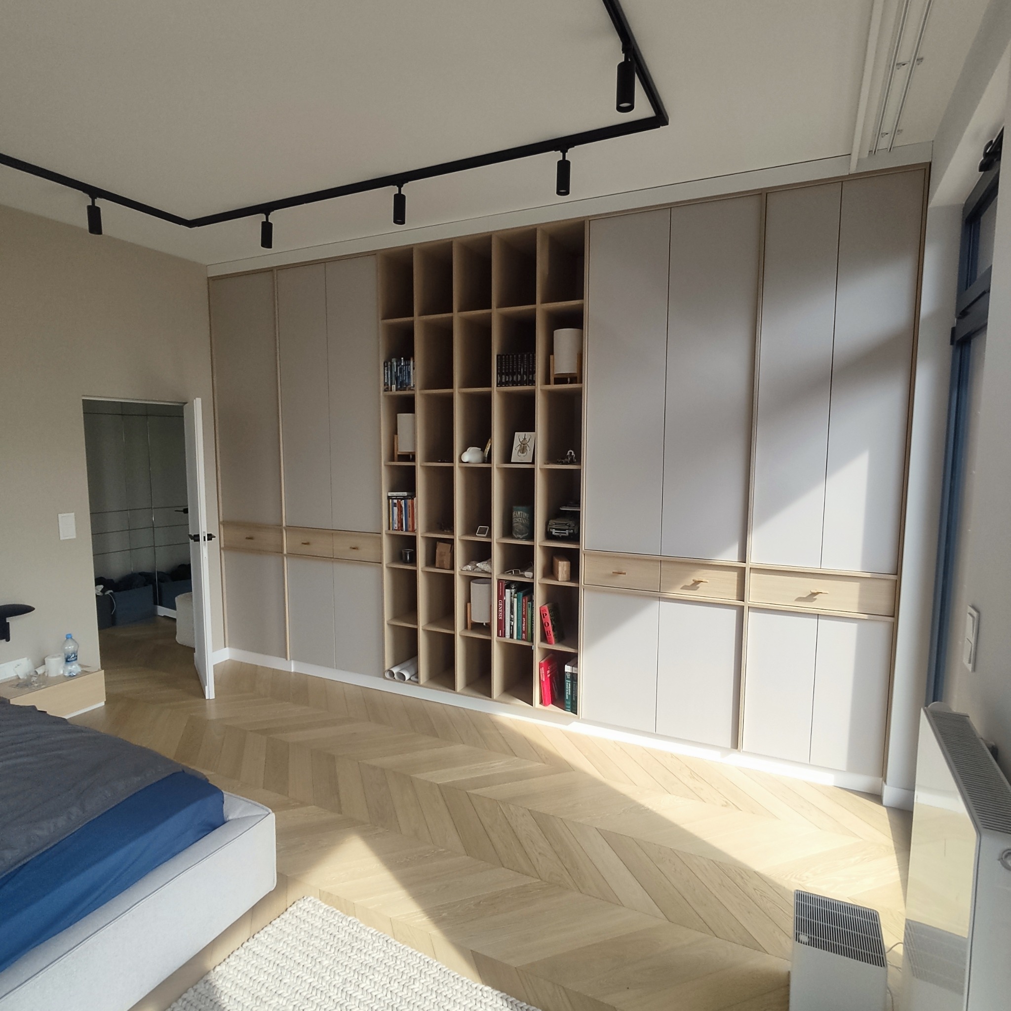 Duża szafa wnękowa z otwartymi półkami na książki i dekoracje, w jasnym pokoju z parkietem ułożonym w jodełkę i czarnym oświetleniem szynowym.