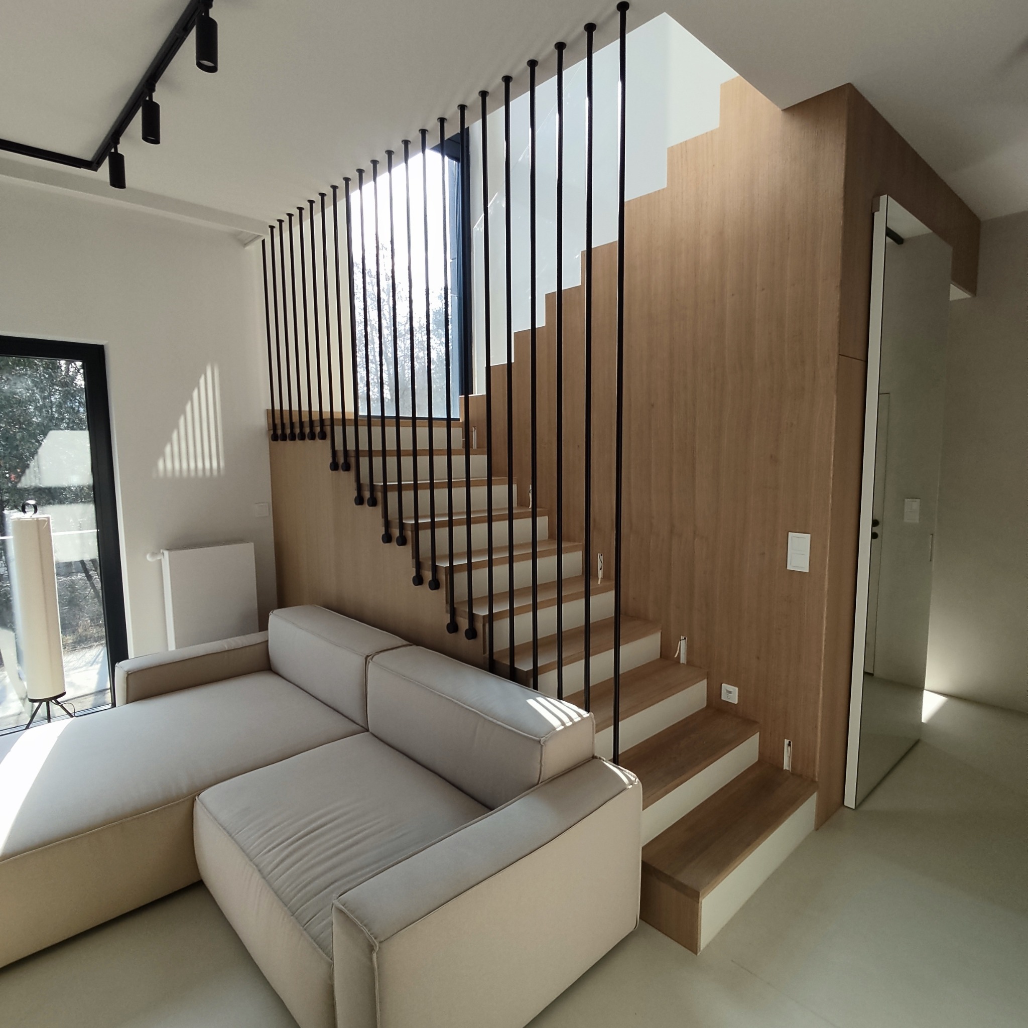 Nowoczesne schody drewniane z czarną balustradą sznurkową, wkomponowane w jasne wnętrze z sofą. Minimalistyczny design, naturalne światło, widok na zewnątrz.