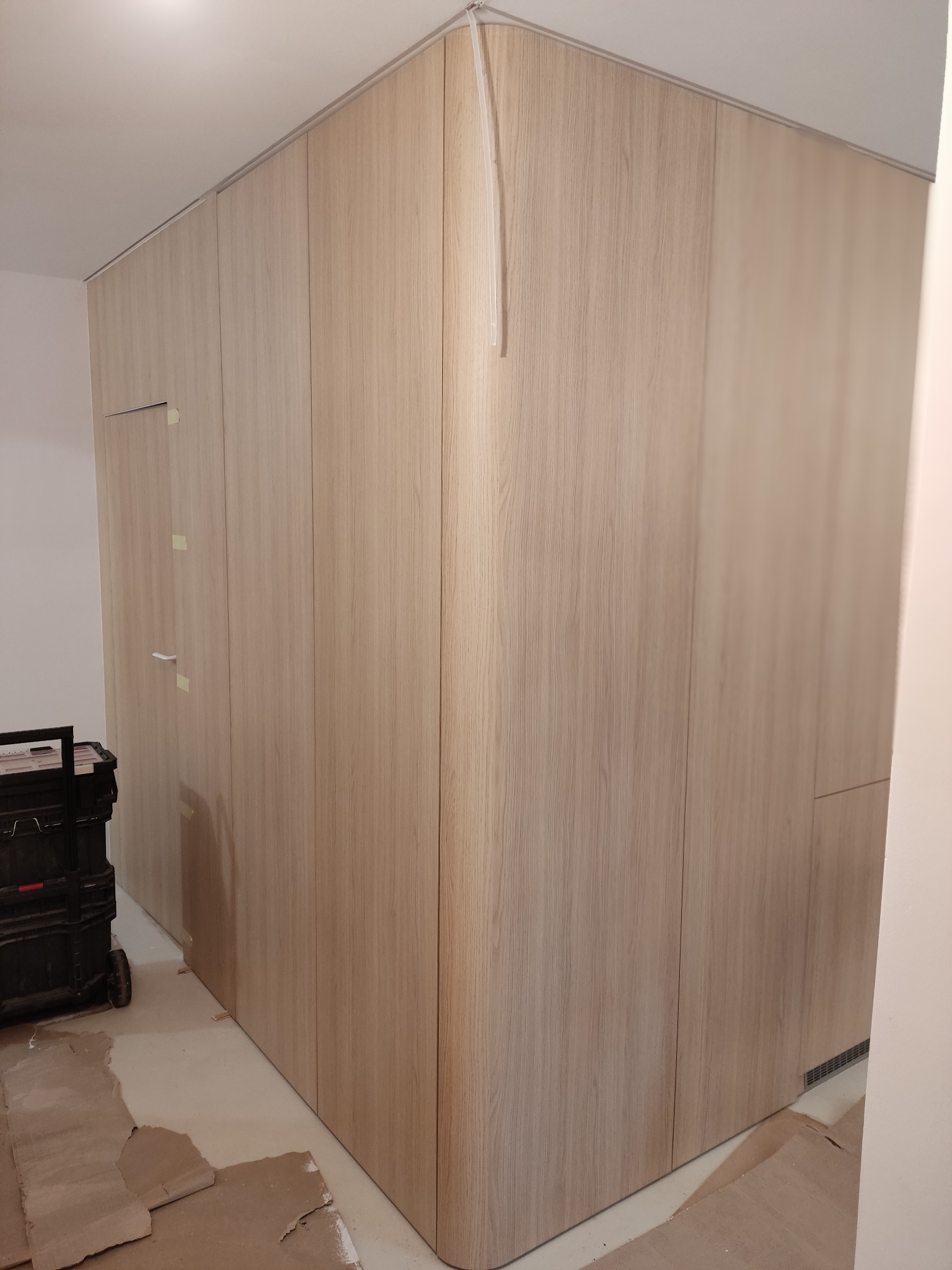 Jasna, drewniana szafa wnękowa z zaokrąglonym narożnikiem, widoczna skrzynka narzędziowa i zabezpieczona podłoga, sugerujące montaż w trakcie realizacji.