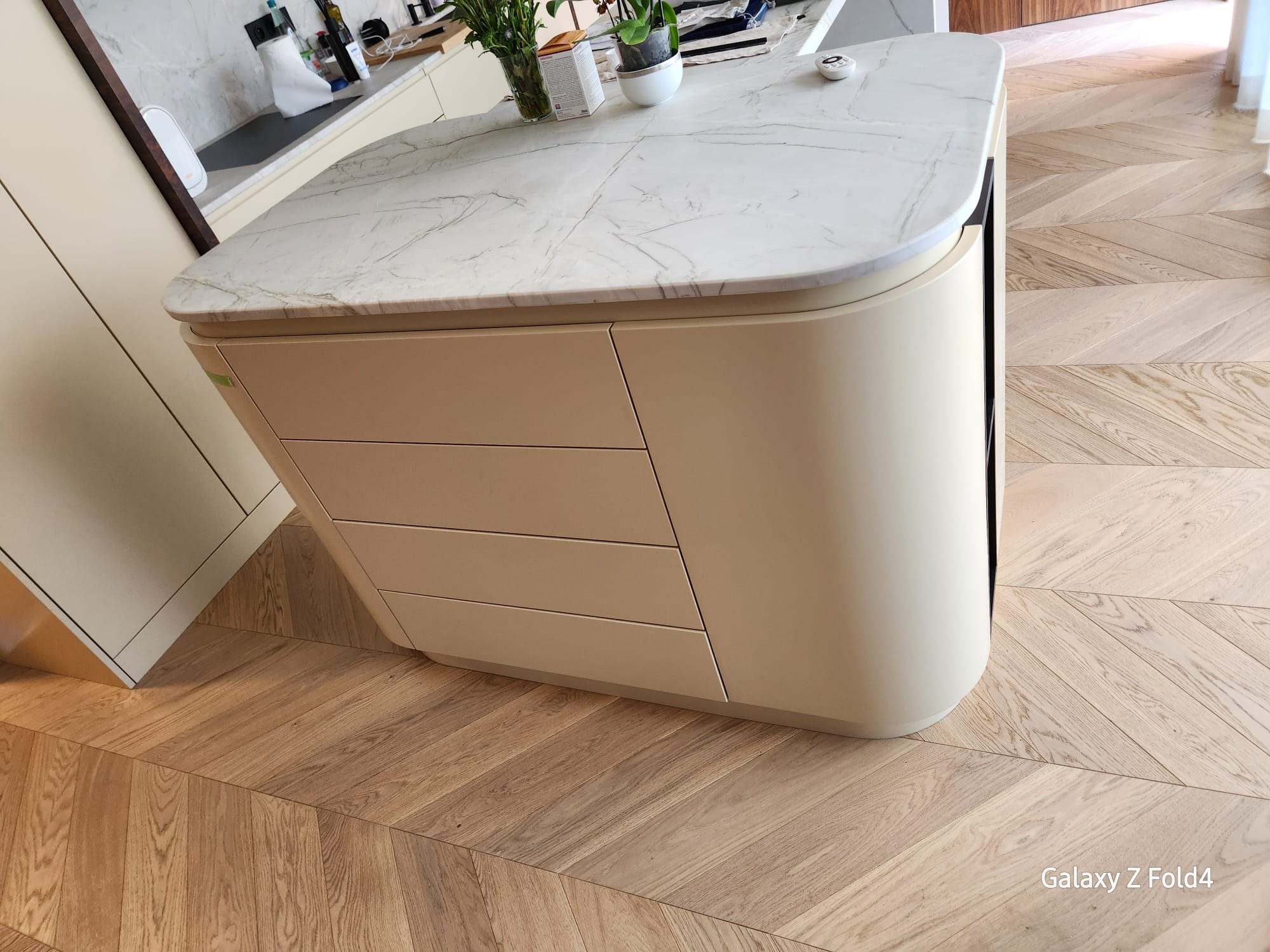 Beżowa wyspa kuchenna z marmurowym blatem na parkiecie w jodełkę. Za wyspą widoczne inne elementy zabudowy kuchennej i rośliny doniczkowe.