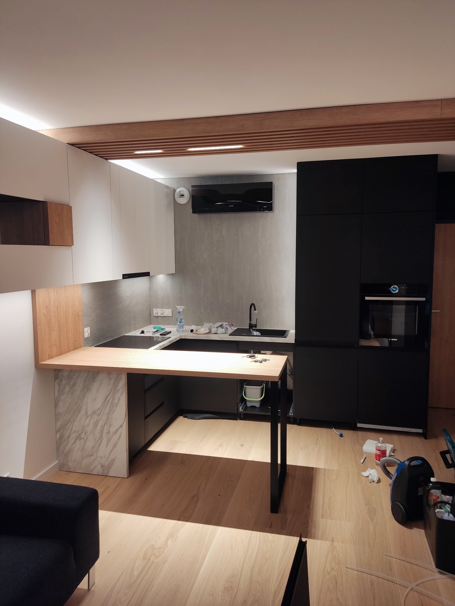 Nowoczesna kuchnia z białymi i czarnymi szafkami, drewnianym blatem i marmurowym wykończeniem. Widoczne elementy wyposażenia i oświetlenie podszafkowe.