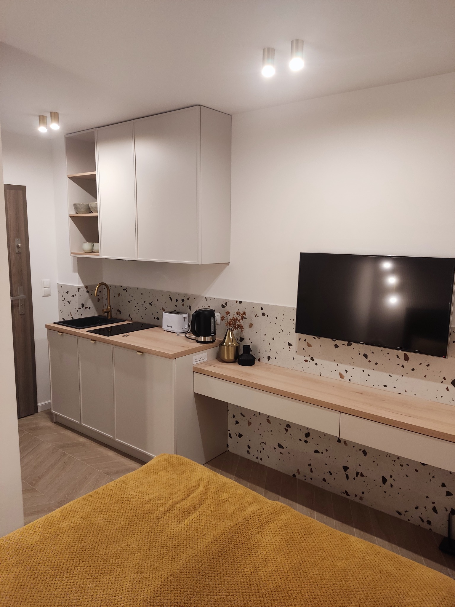 Funkcjonalna kuchnia na wymiar w małym mieszkaniu: jasne szafki, blat z drewna, złota armatura, mozaika na ścianie i telewizor. Kompaktowe rozwiązanie.