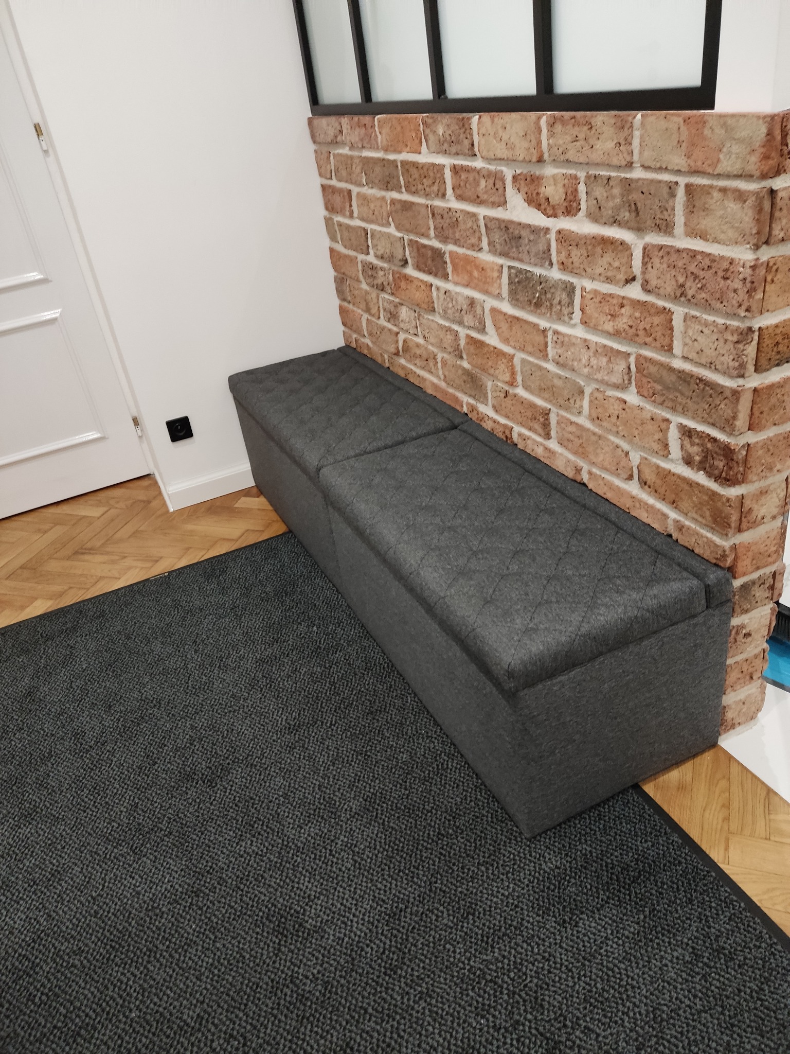 Szara, pikowana skrzynia na pościel w aranżacji wnętrza w Krakowie. Mebel stoi przy ścianie z cegły, na ciemnym dywanie. Widoczna podłoga z parkietu i fragment drzwi.