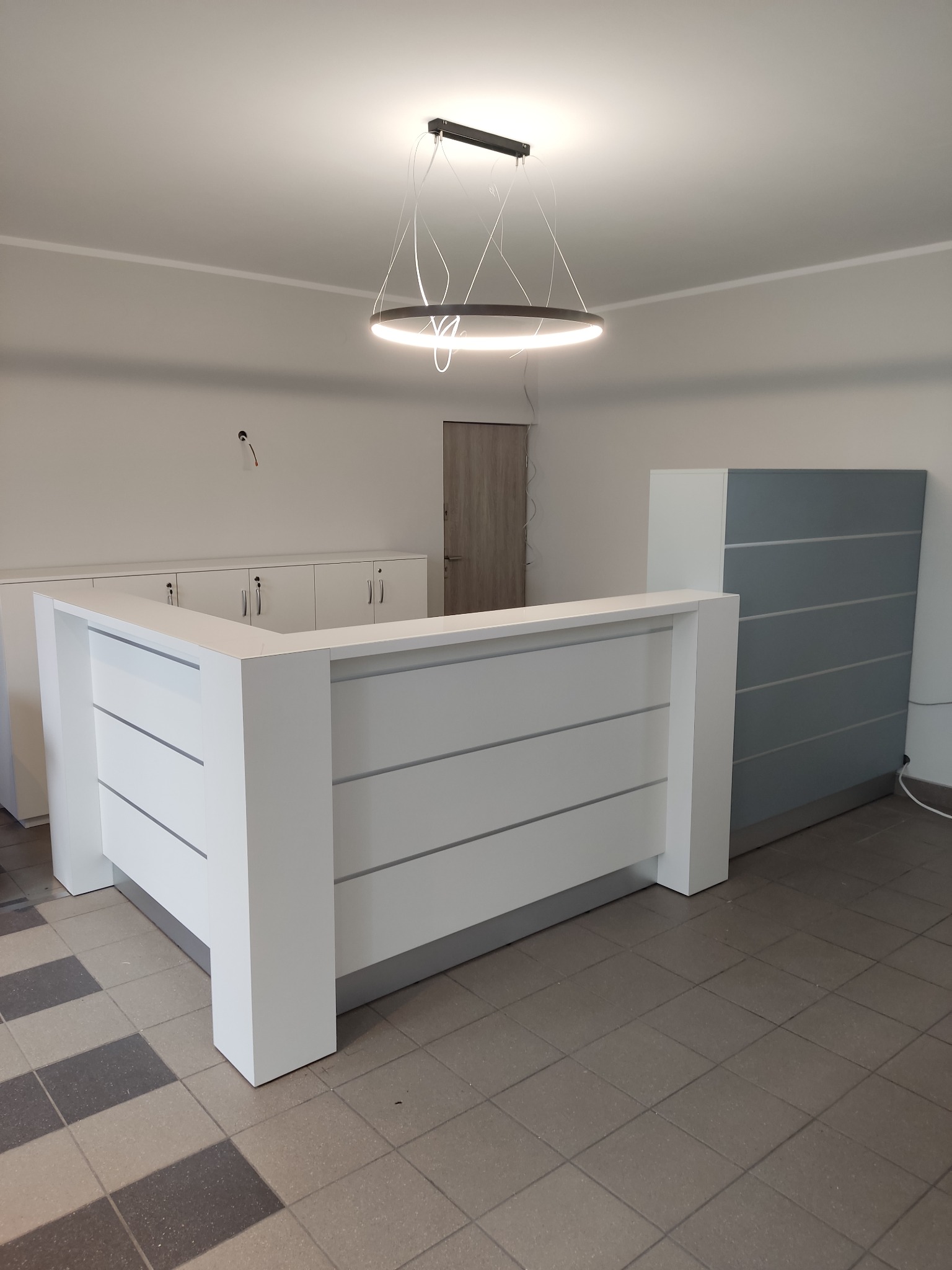 Biała lada recepcyjna z poziomymi panelami, oświetlona nowoczesną lampą. W tle szafki zamykane na klucz i szara szafa. Całość w minimalistycznym, nowoczesnym stylu.