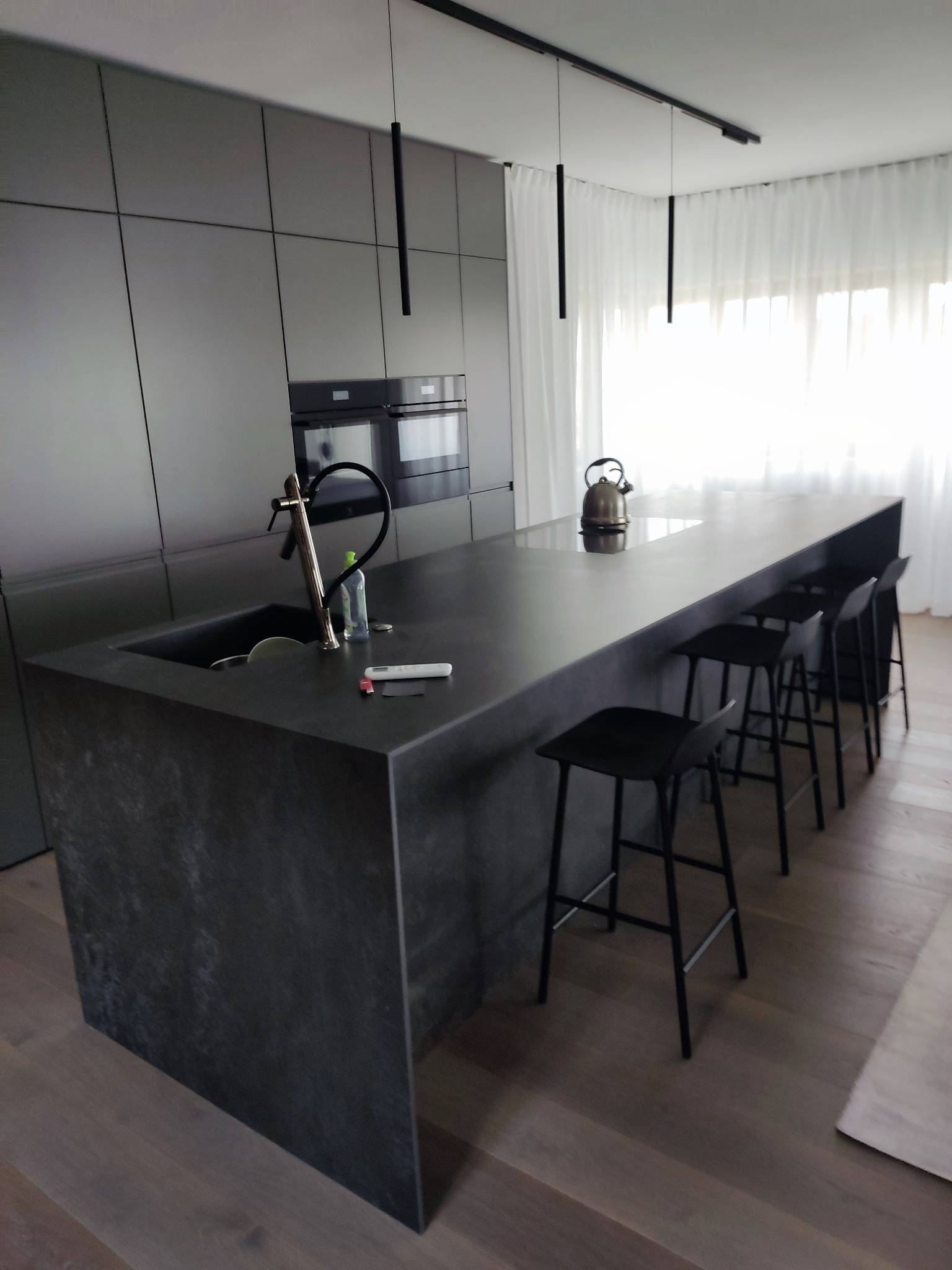 Nowoczesna kuchnia z wyspą i hokerami. Ciemnoszara zabudowa meblowa, minimalistyczne lampy wiszące. Na blacie czajnik i akcesoria kuchenne. Prosty, elegancki design.