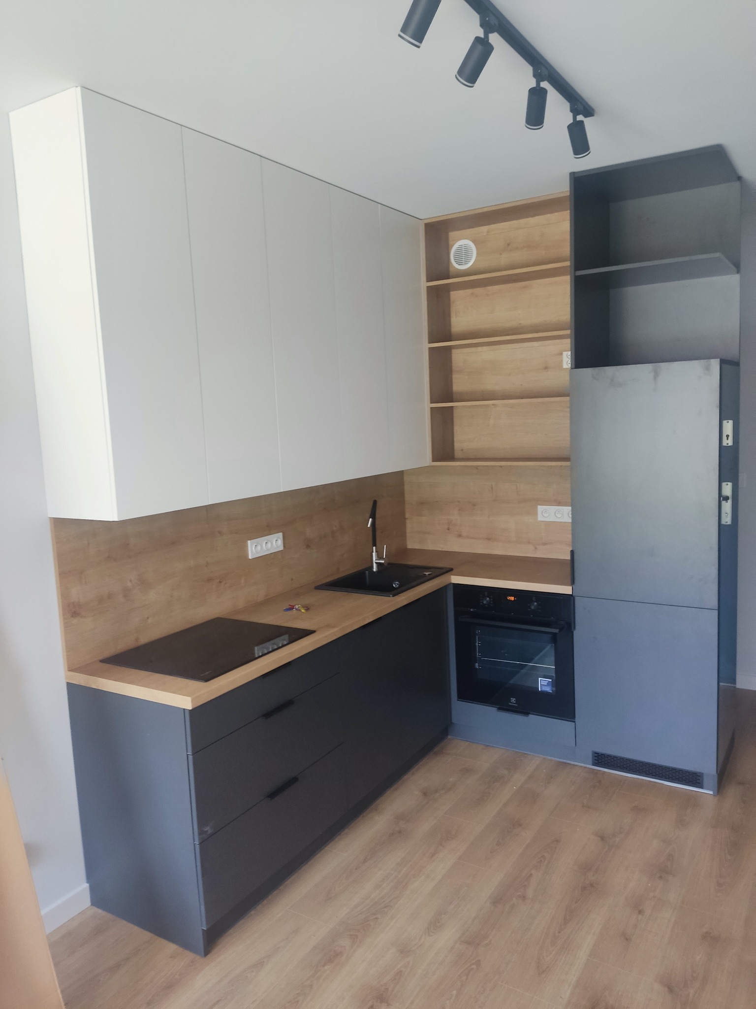 Nowoczesna kuchnia z szarymi szafkami dolnymi i białymi górnymi, drewnianym blatem oraz otwartymi półkami z jasnego drewna, oświetlona punktowymi lampami.