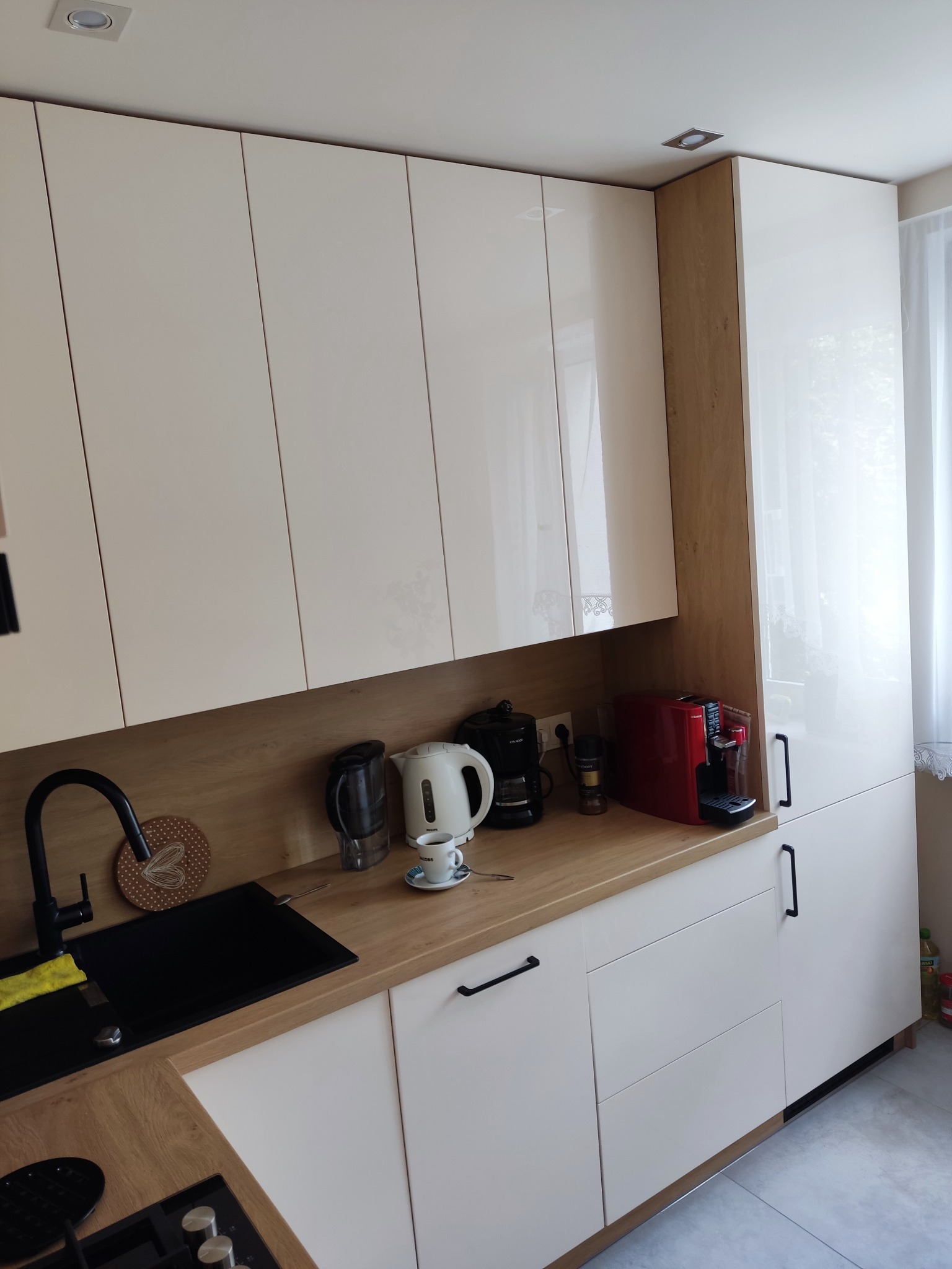 Biało-kremowa kuchnia z połyskiem i drewnianym blatem. Widoczny czarny zlew, ekspresy do kawy i czarne, podłużne uchwyty. Górne szafki sięgają sufitu.