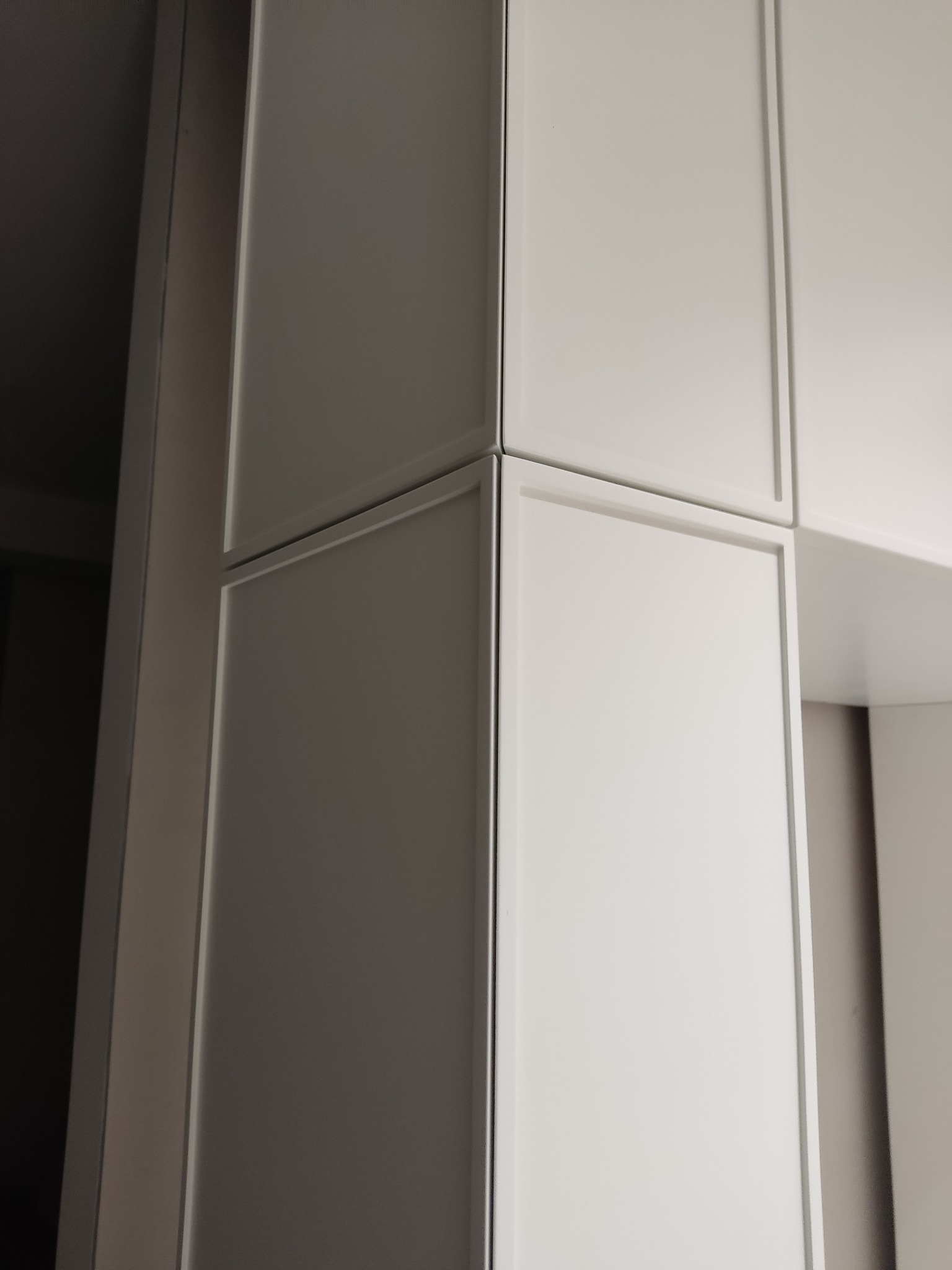Białe, matowe fronty szafy wnękowej z delikatnym frezem, widoczne pod kątem. Minimalistyczny design, subtelne oświetlenie, fragment wnętrza.