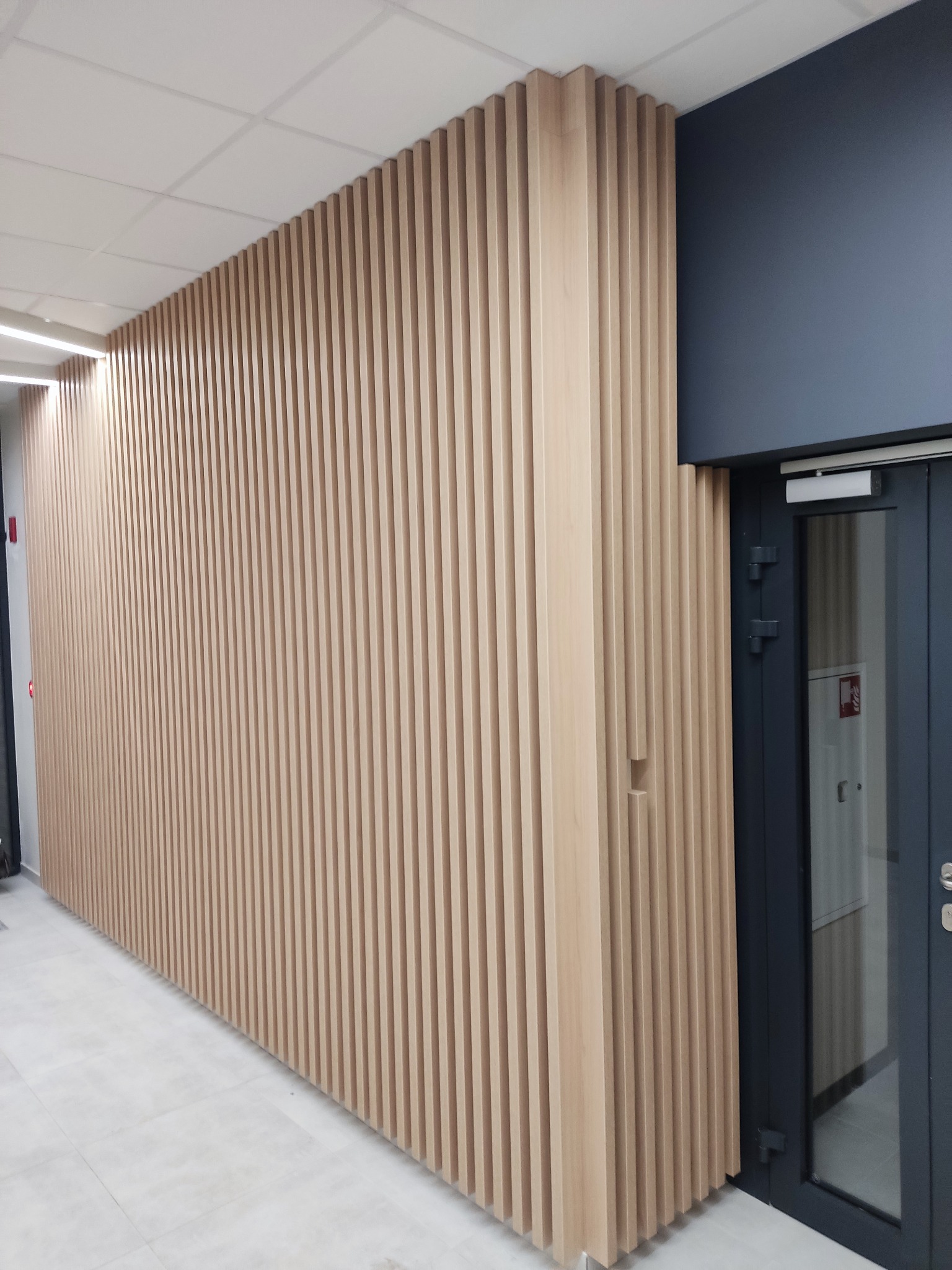 Drewniana ściana lamelowa w korytarzu biurowym, pionowe listwy w jasnym odcieniu drewna, widoczna część drzwi wejściowych i sufit podwieszany.
