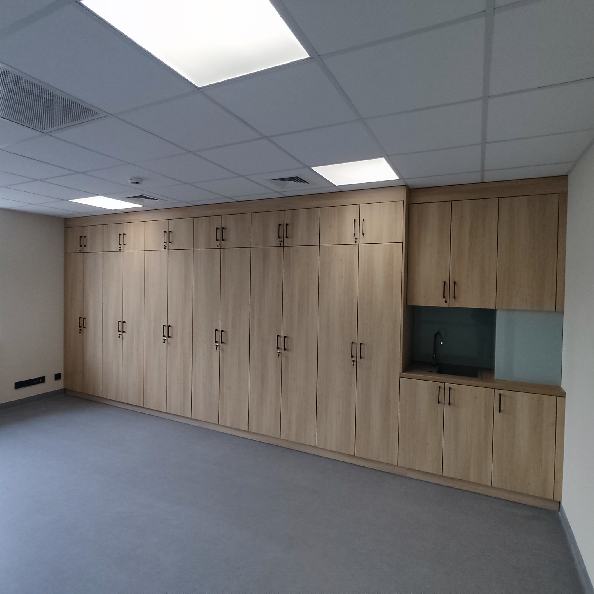 Długa szafa z jasnego drewna z czarnymi uchwytami, wbudowana umywalka z baterią. Nowoczesne wnętrze z szarą podłogą i oświetleniem sufitowym.