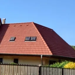 Czerwony dach z dachówką karpiówką i dwoma oknami dachowymi, widziany z poziomu gruntu za białym płotem, w tle błękitne niebo.