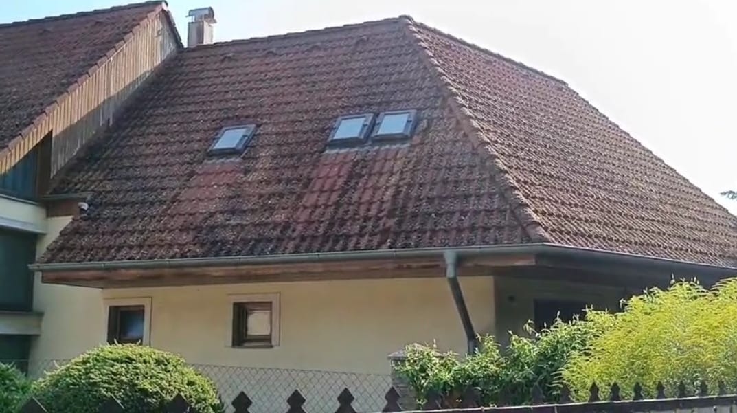 Czerwony dach z dachówką ceramiczną, pokryty mchem, z trzema oknami dachowymi, widoczne rynny i fragment elewacji domu.