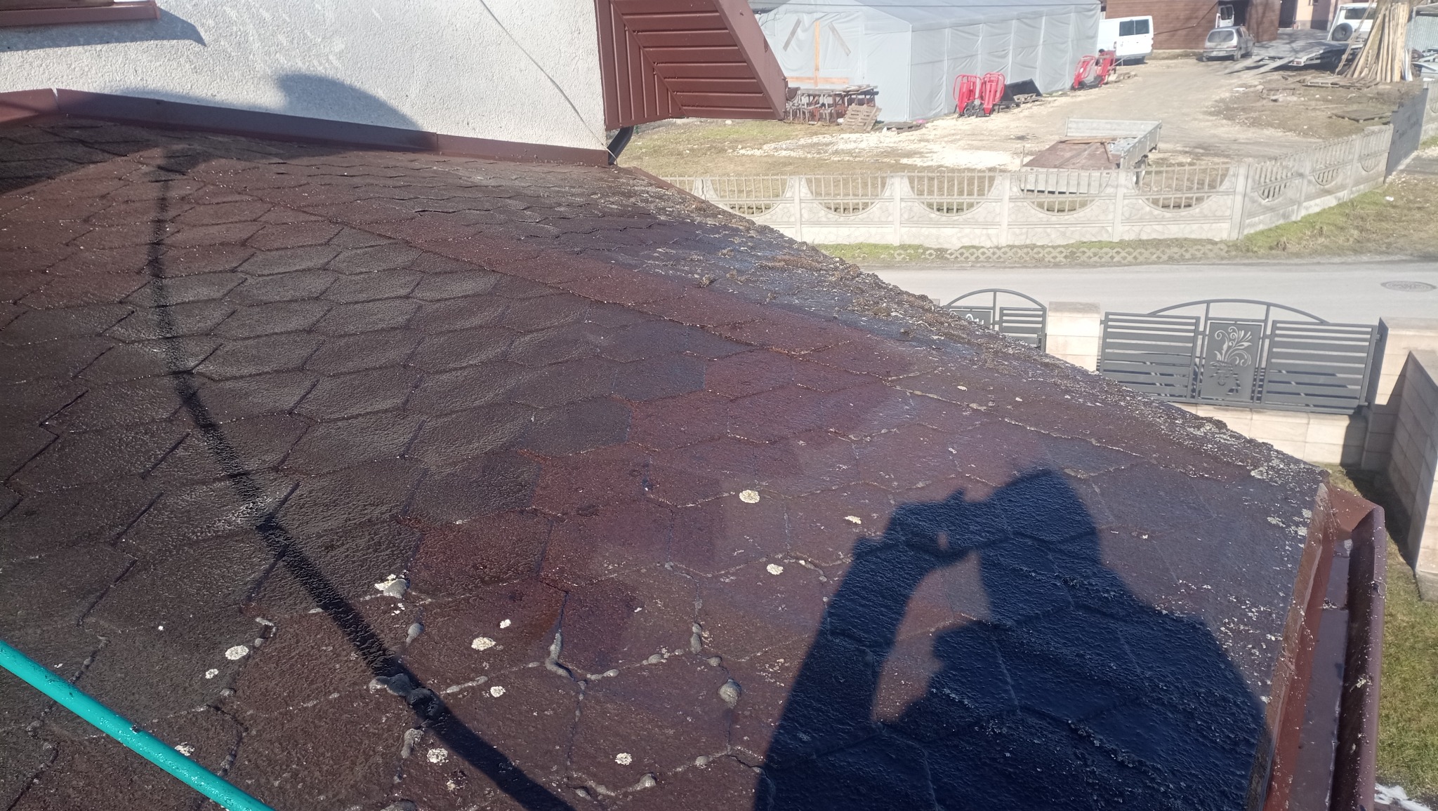 Mokry, brązowy dach z gontów bitumicznych po czyszczeniu, z widocznym cieniem osoby i fragmentem węża. W tle brama i zabudowania.