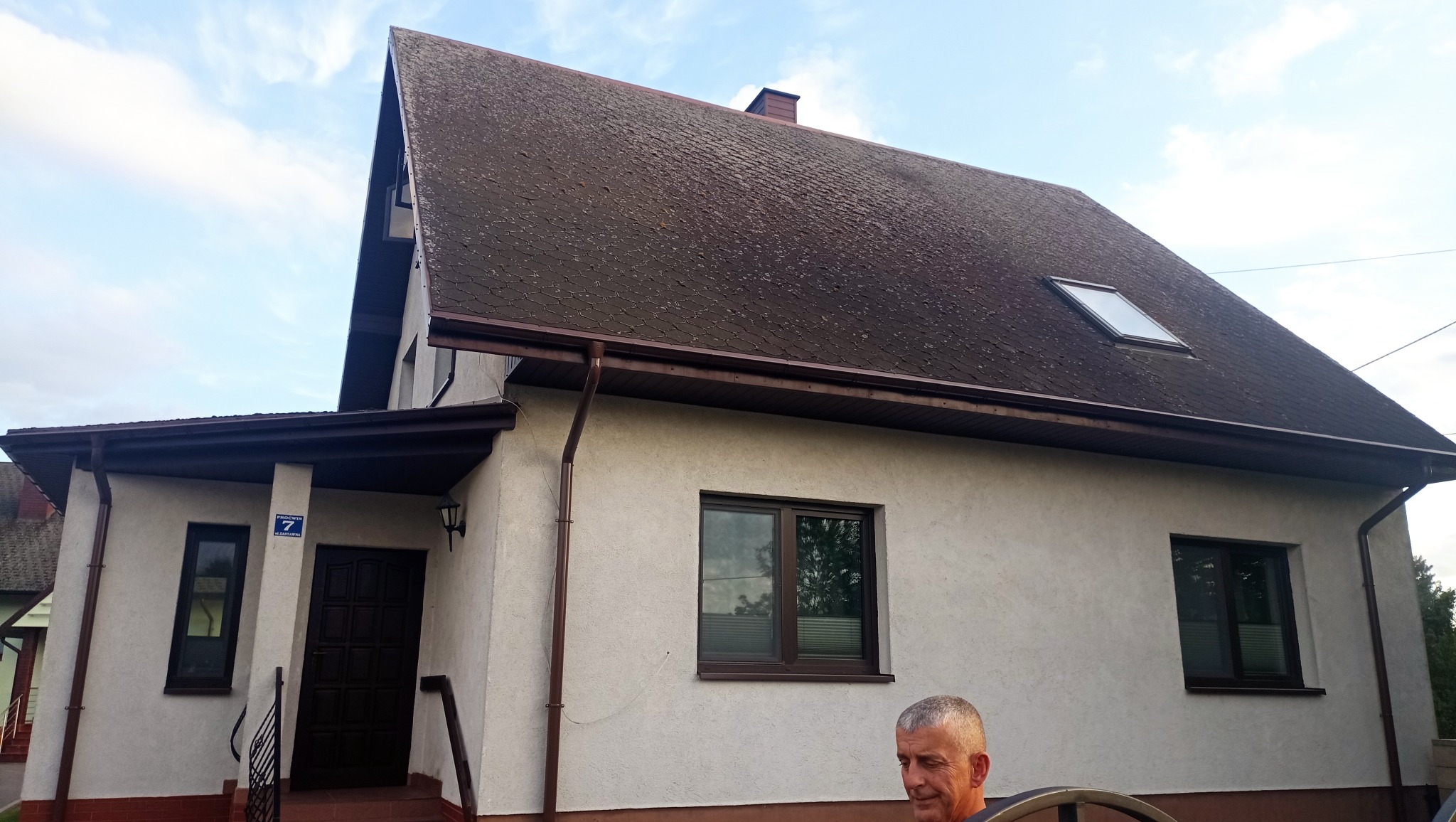 Dom jednorodzinny z ciemnym, zabrudzonym dachem i jasną elewacją. Widoczne okna, rynny i fragment osoby w dolnej części kadru. Dach pokryty mchem i zabrudzeniami.