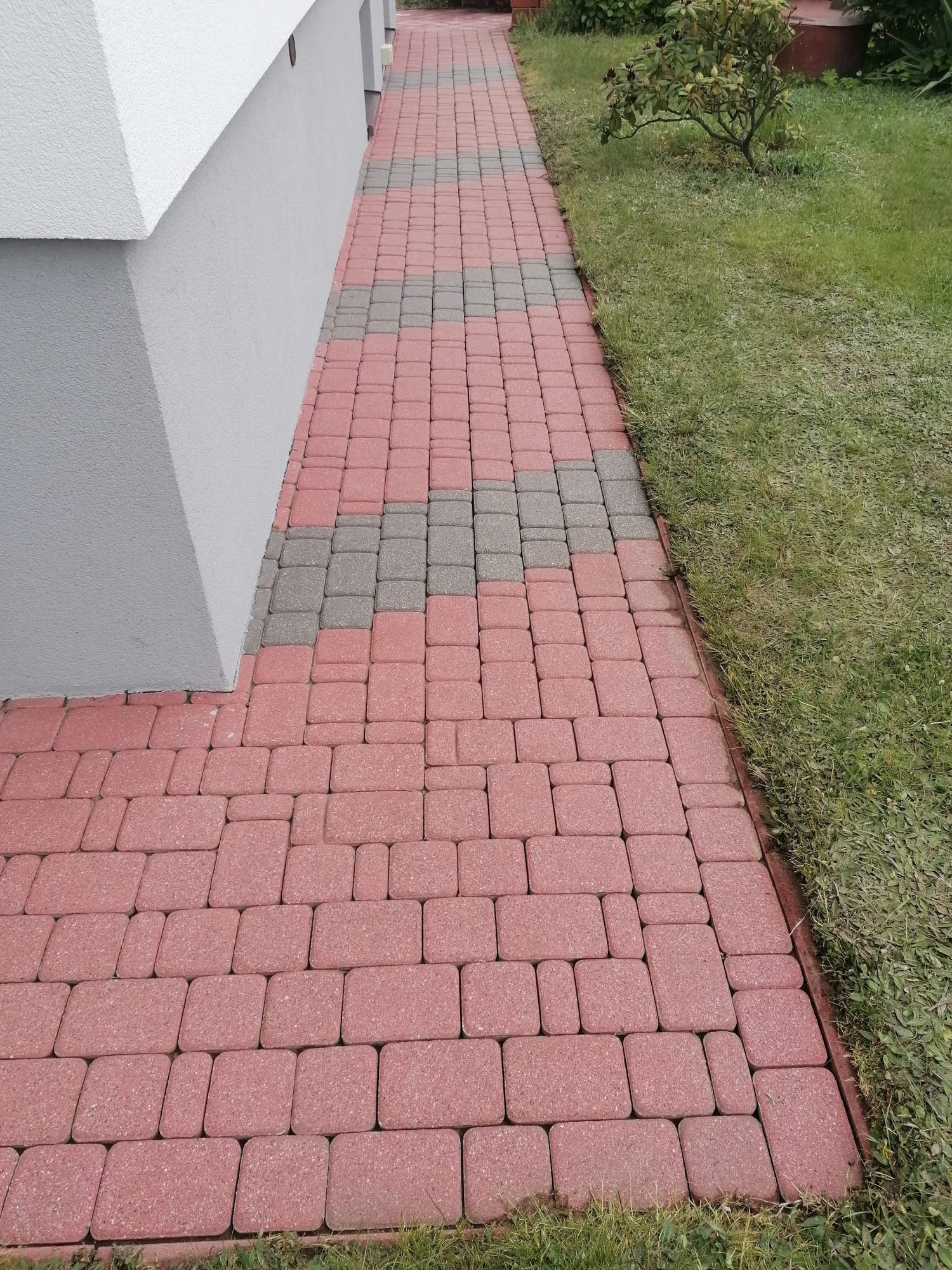 Ułożona, czerwono-szara kostka brukowa tworząca ścieżkę obok budynku i trawnika. Widok z góry, perspektywa zbieżna. Kostka ułożona w regularny wzór.