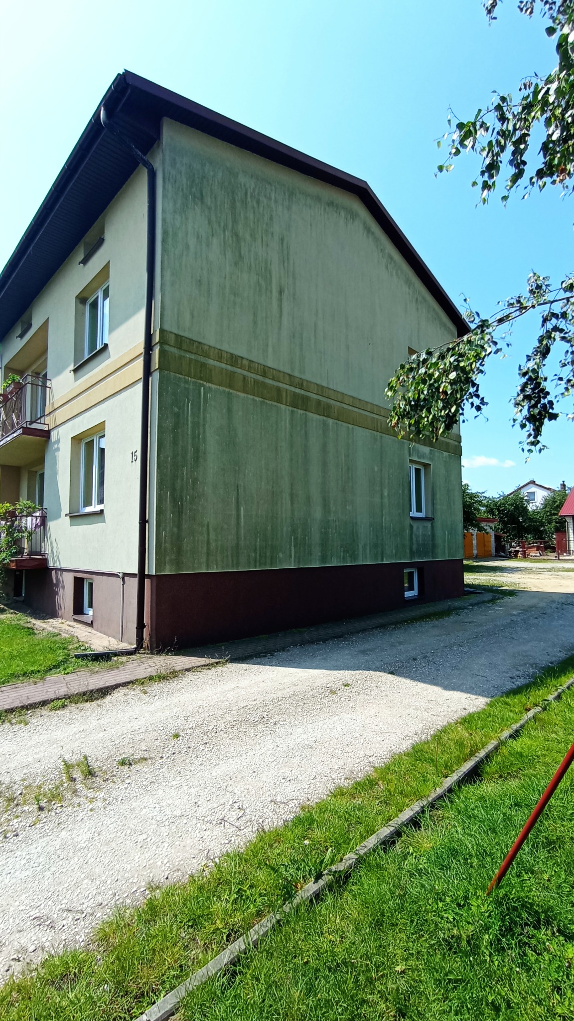 Zaniedbana elewacja dwupiętrowego budynku z widocznymi zabrudzeniami i zaciekami, z numerem 15, na tle błękitnego nieba i zieleni drzew.