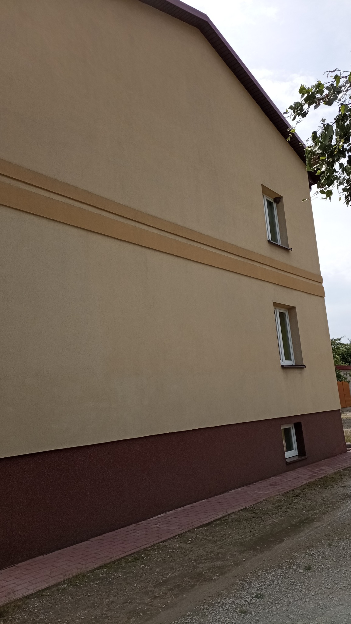 Dwukondygnacyjny dom z beżową elewacją, brązowym cokołem i trzema oknami. Widok z dołu, z fragmentem dachu i zielenią w tle. Ścieżka z kostki brukowej.