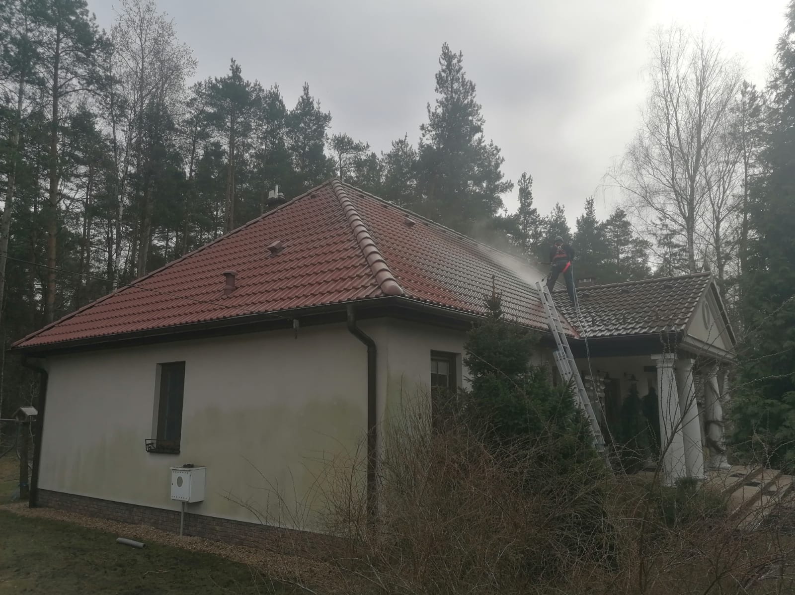 Pracownik na drabinie czyści dach domu z czerwonej dachówki za pomocą myjki ciśnieniowej. Część dachu jest już umyta, widoczna różnica w kolorze.