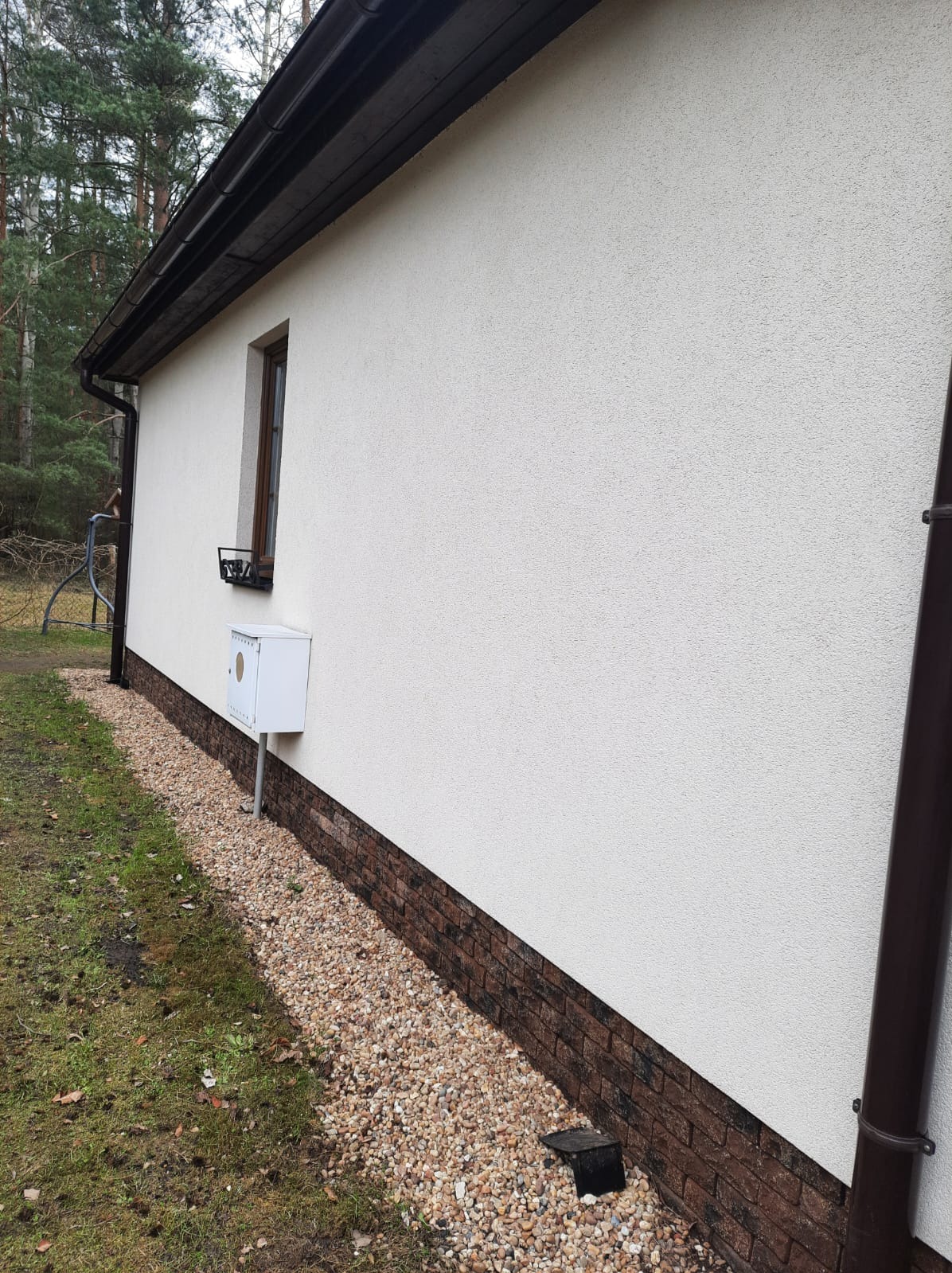 Boczny widok domu z jasną elewacją, brązowym cokołem z cegły i rynną. Widoczne okno z ozdobną kratką i skrzynka gazowa. Tło stanowią drzewa.