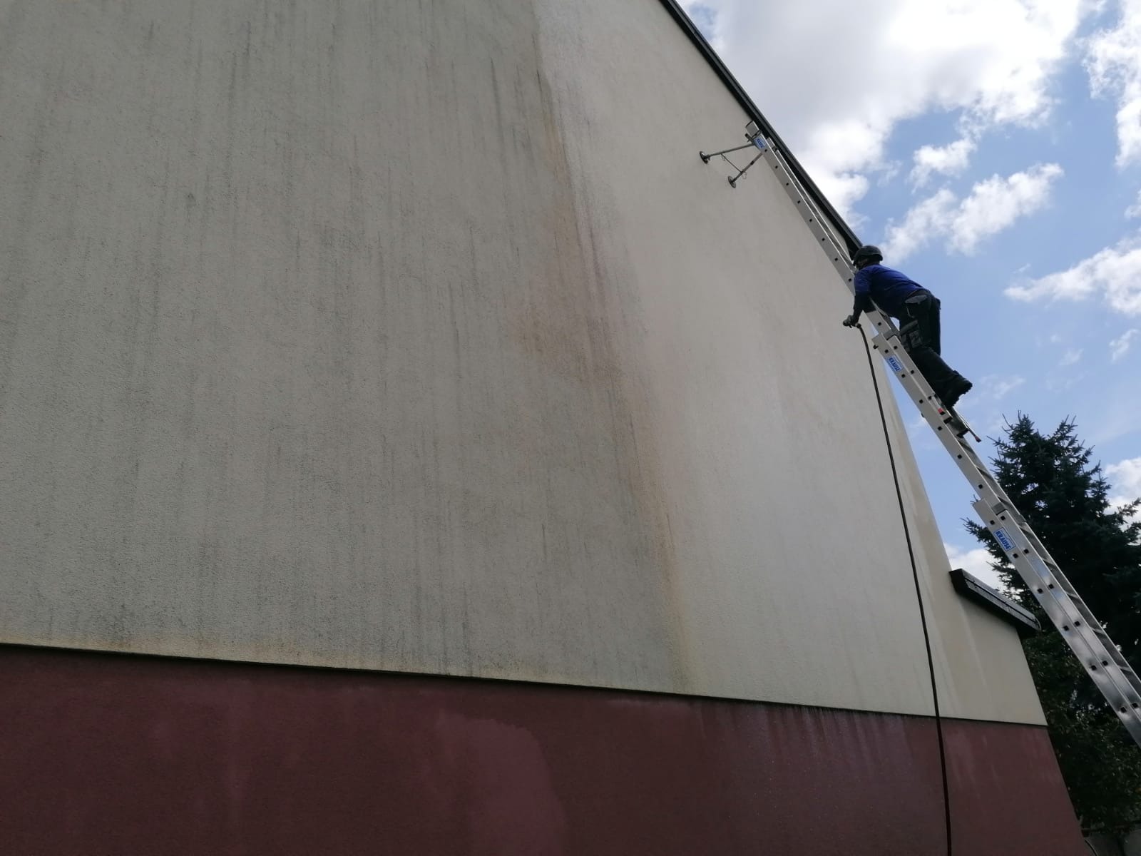 Pracownik na drabinie czyści elewację budynku. Widoczna różnica w czystości ściany, od dołu ciemniejsza, góra jaśniejsza. Błękitne niebo w tle.