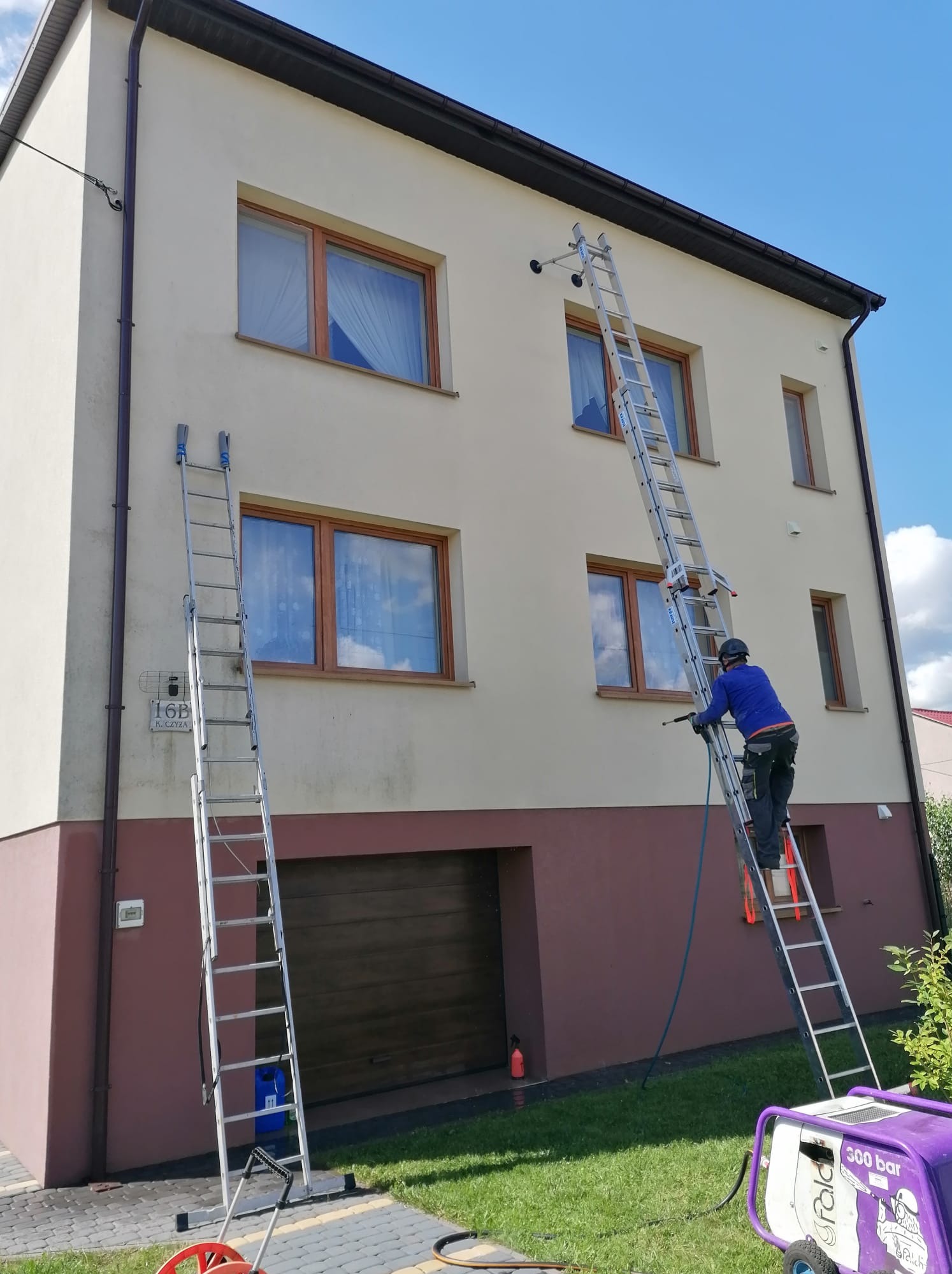 Pracownik na drabinie, w kasku, z lancą ciśnieniową, czyści elewację dwukondygnacyjnego domu. Widoczny sprzęt ciśnieniowy na trawniku i druga drabina oparta o ścianę.