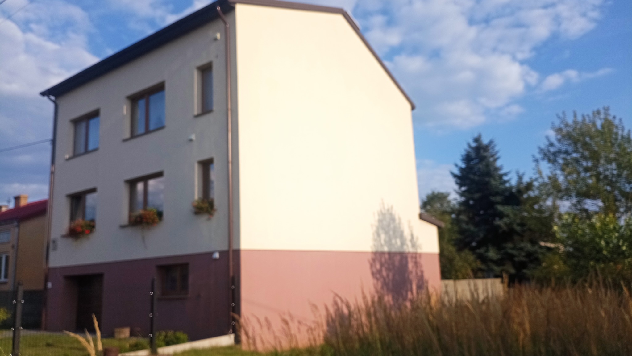 Dwukondygnacyjny dom z jasną elewacją i brązowym cokołem, widoczny z perspektywy trawnika. Okna z kwiatami, błękitne niebo w tle. Częściowo widoczny budynek sąsiedni.